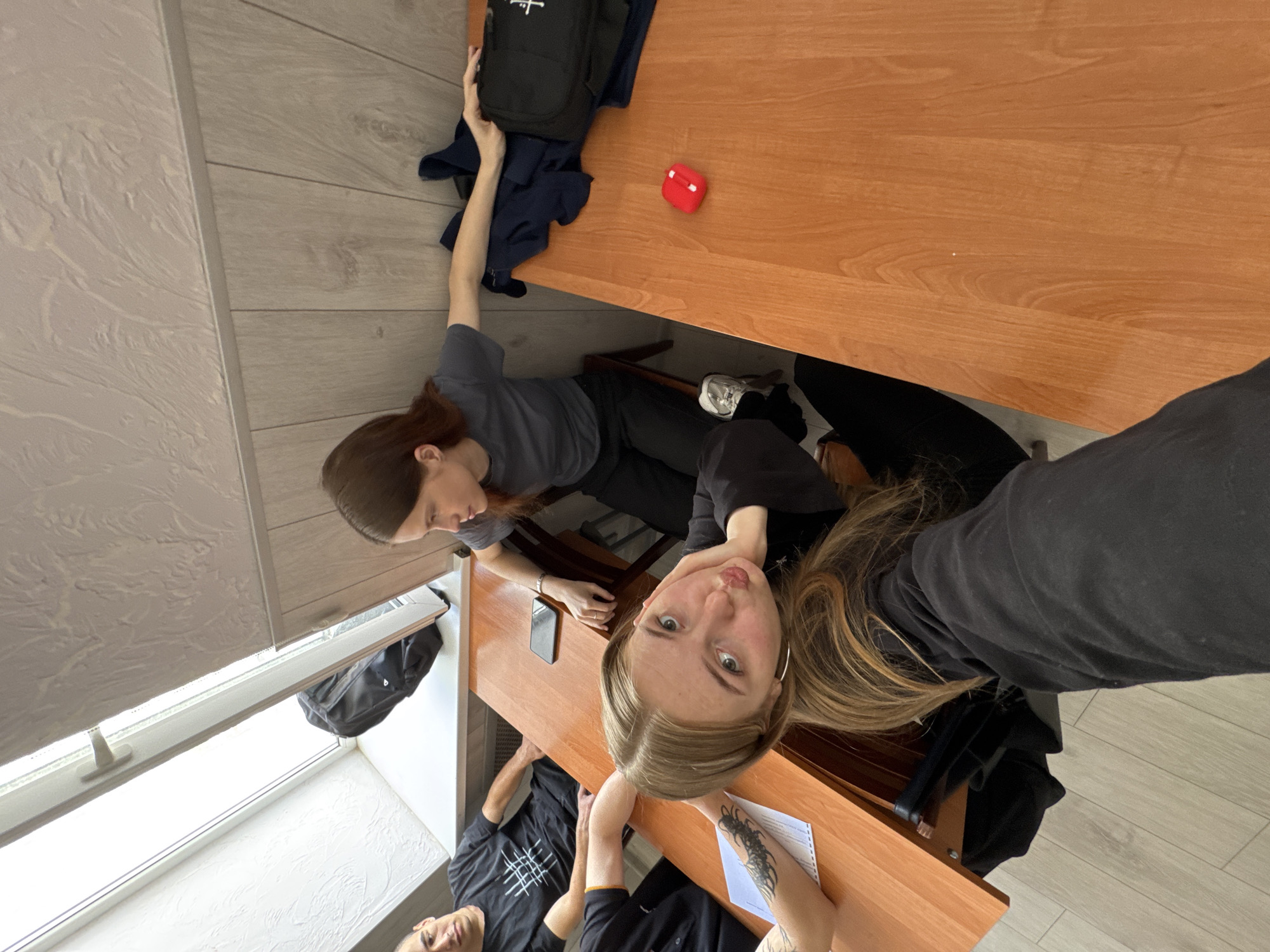 Two women and a man are in a room. The women are seated at desks and the man is seated at the table. One woman has light brown hair, while the other woman has dark brown hair. All three are wearing black clothing. A bookbag, red object, and water bottle are visible on a desk. A window is visible with white blinds. The specific location is not identifiable from the image.