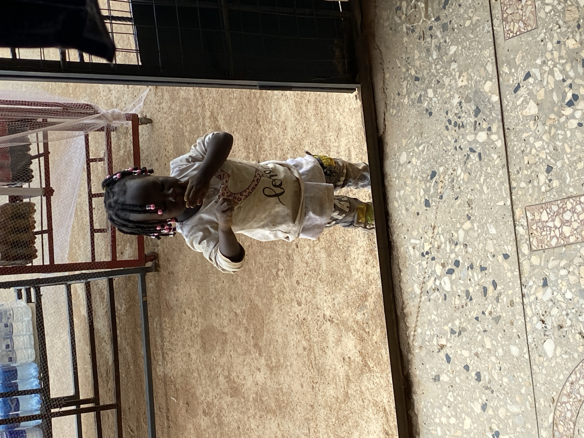 A daytime photograph captures a young child, likely a girl, standing in what appears to be a doorway or transition area in Bukuru, Nigeria. The child has dark braided hair adorned with pink beads and is wearing a light-colored t-shirt with partial dark cursive text, possibly "Lover," and patterned trousers. She stands on a sandy or dirt floor, with her right hand raised towards her face, conveying a shy or contemplative expression.
Behind the child, on the left, there's a metal frame that could be a bed or bunk bed, partially covered by a white, translucent mosquito net. Further to the left, a metal rack holds several stacked large plastic bottles, presumably containing water. The right side of the image reveals a contrasting floor of speckled concrete or terrazzo, suggesting a more finished interior or patio area. The overall scene is brightly lit, indicating daylight. The details like the mosquito net, stacked water bottles, and varied flooring textures provide specific location cues consistent with a practical, lived-in environment in the region.