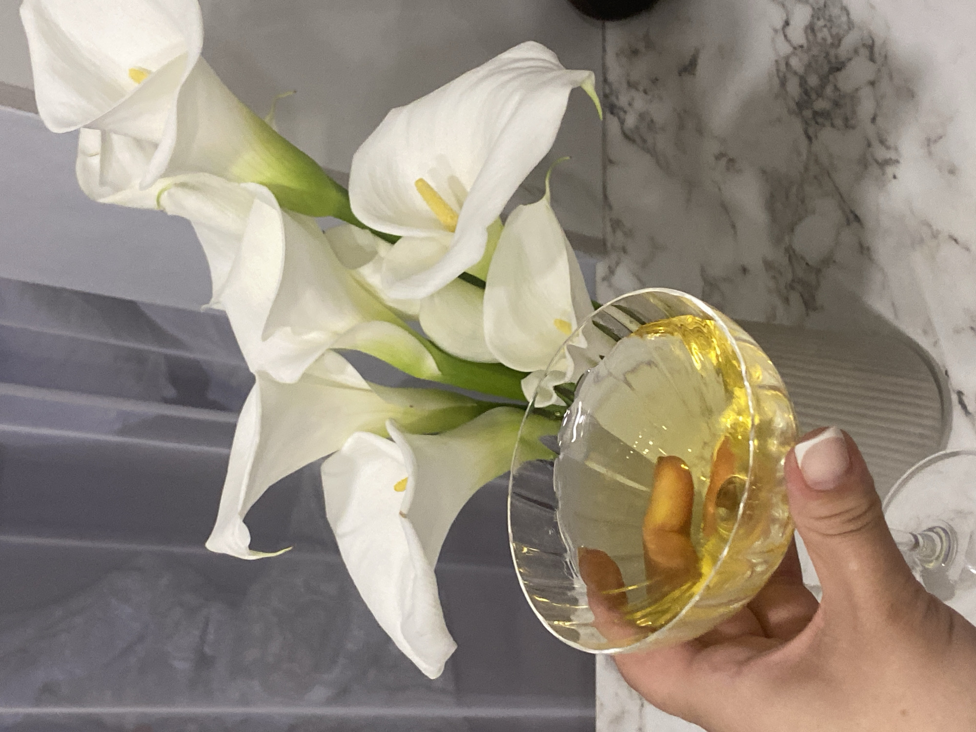 The image depicts an indoor scene in the city of Sochi, Russia. In the foreground, a light-skinned hand, featuring a French manicure on the visible thumb and index finger, grasps the stem of a clear glass. The glass, which appears to be a coupe or wide-rimmed cocktail glass, has a fluted pattern on its bowl and contains a yellowish liquid with two small, rectangular, orange-brown pieces submerged within it. Positioned centrally in the mid-ground, a bouquet of approximately six white calla lilies, each with a bright yellow spadix, is arranged in a light grey, vertically ribbed cylindrical vase. The entire display rests on a polished white marble surface distinguished by intricate grey and black veining. The background is composed of muted grey-blue or charcoal-colored curtains or drapes, which exhibit vertical folds and a soft, diffused quality.