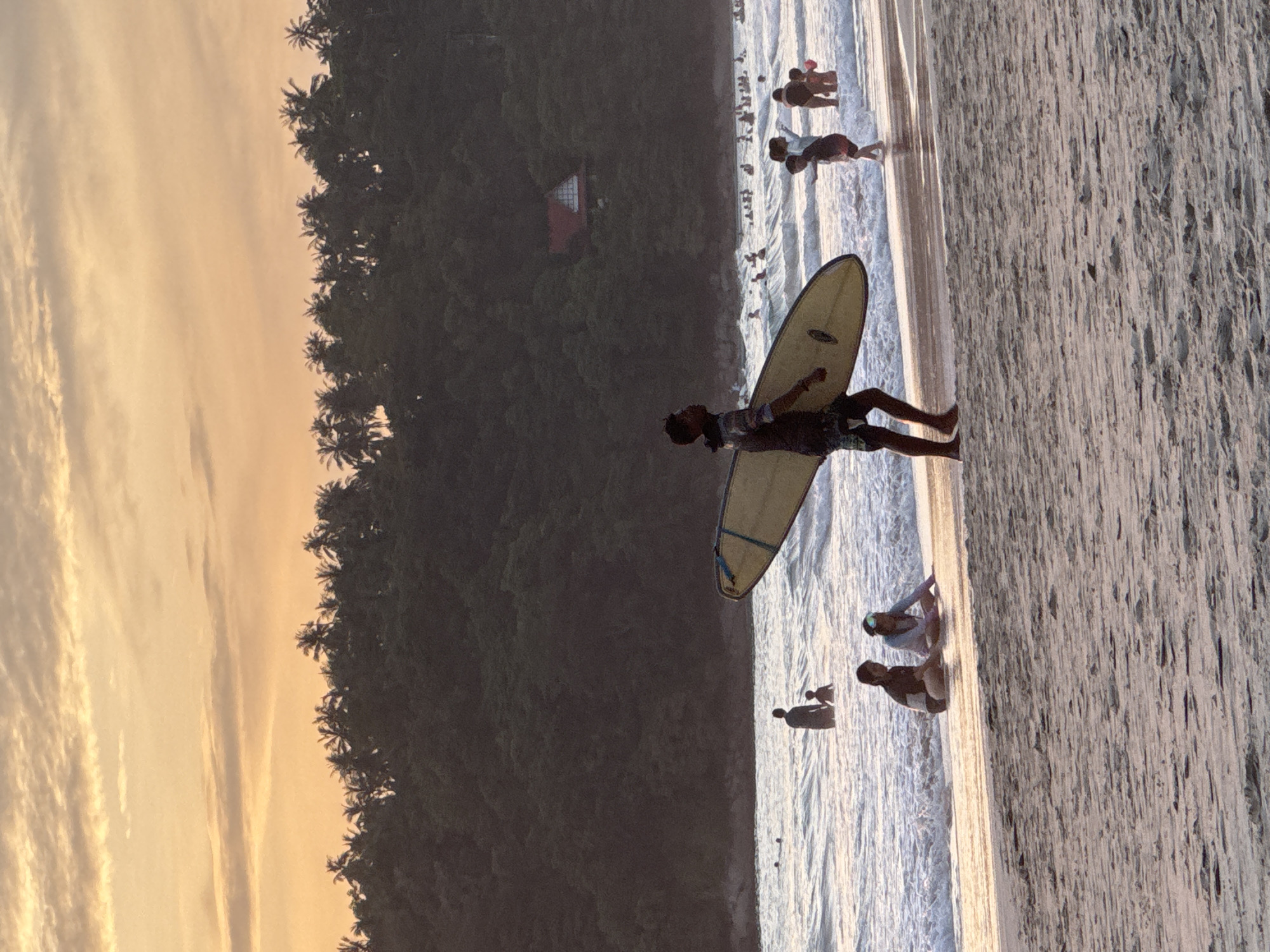 A man carries a surfboard along Karon Beach in Karon, Thailand, during a sunset. The sky is filled with pastel colors and clouds, and a dense line of palm trees borders the lush, tree-covered hills that rise behind the beach. The ocean is calm with gentle waves, and several people are swimming and playing in the water. On the sandy shore, two women are sitting, and further down the beach, a group of children are interacting in the shallow water. The sand is wet near the water's edge and dry further up the beach, showing footprints and indentations.
