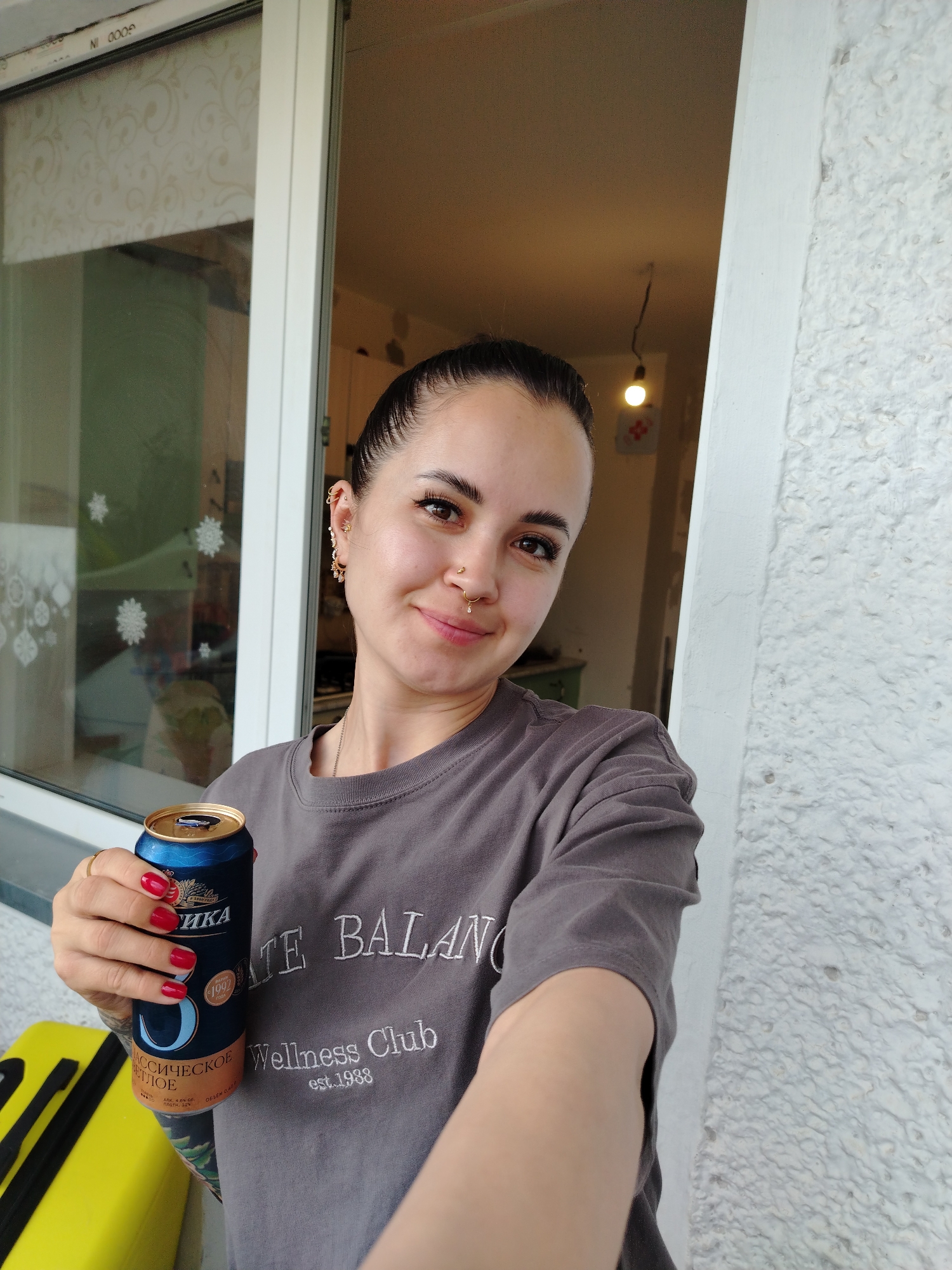 A woman with dark hair pulled back and multiple piercings is holding a can of beer in her right hand. She is wearing a gray t-shirt with embroidered text that reads "BALANCE Wellness Club est. 1988". Her nails are painted red. To her left, a yellow suitcase is visible. The background suggests an outdoor setting, possibly a balcony or porch, with a window featuring a decorative pattern and a textured white wall to the right. The scene appears to be in Noviy, Russia, though no specific landmarks are identifiable within the image.