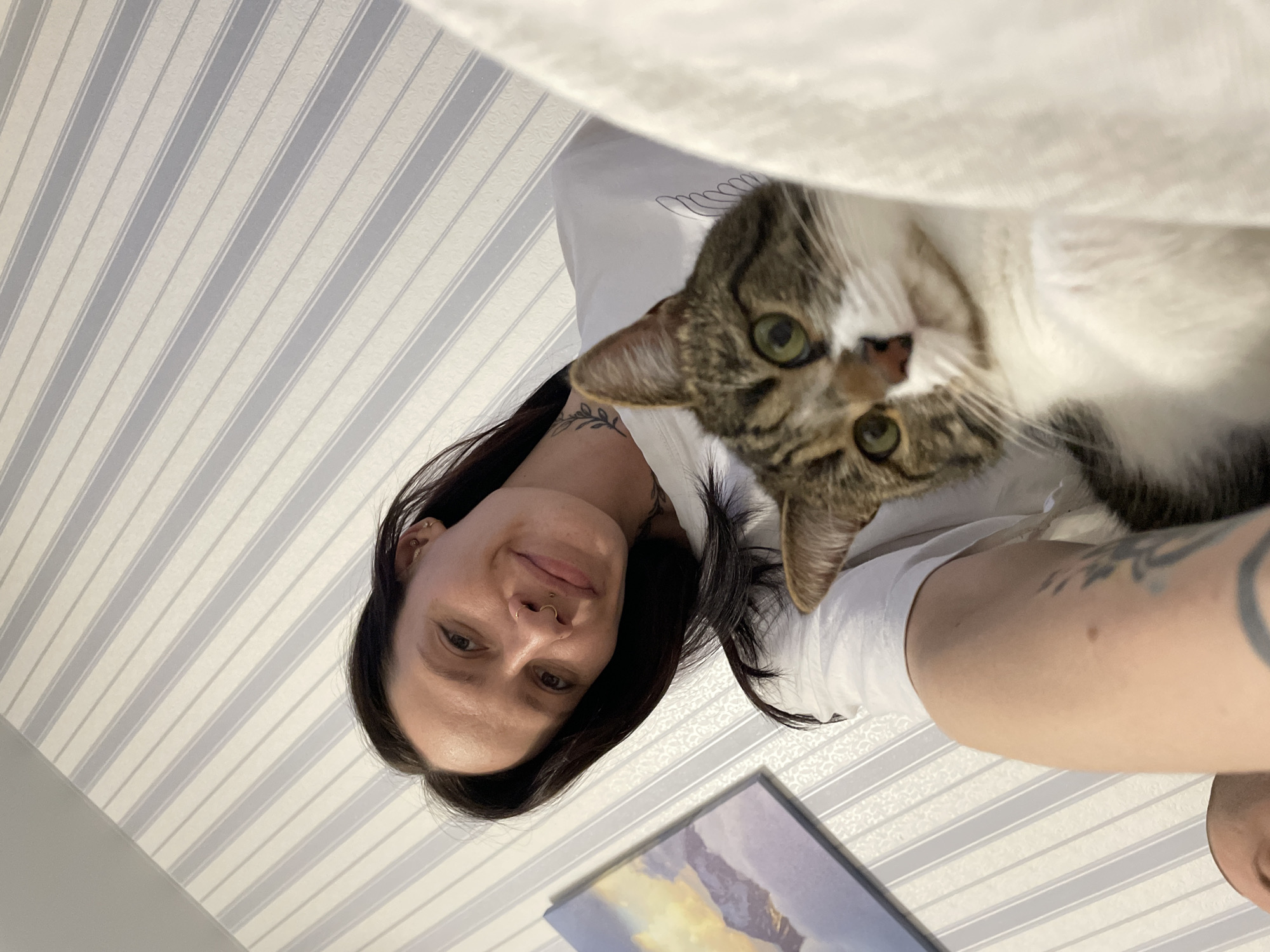 A woman with dark hair and a nose ring is holding a brown and white tabby cat. The woman is smiling and looking at the camera. The cat is also looking at the camera. The background is a striped wall and a framed picture. There is no indication this image was taken in Izhevsk, Russia.