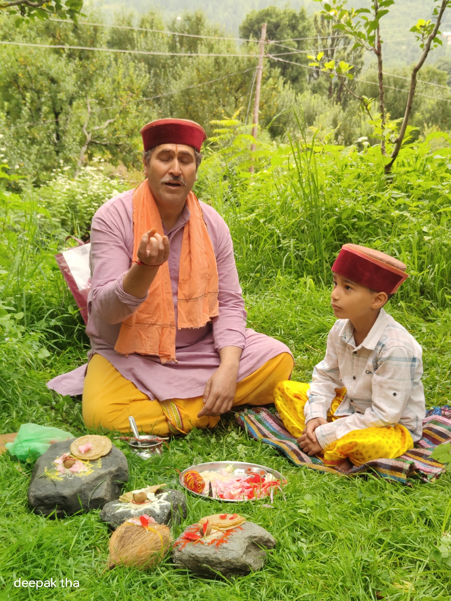 In Vashisht, India, two individuals, a man and a boy, are seated on the grass in a lush, green outdoor environment, likely a garden or hillside. The man, who appears to be of middle age, is wearing a traditional Himachali cap, an orange scarf over a pink kurta, and yellow lowers. He is gesturing with his right hand, as if explaining or performing a ritual. The boy, who is younger, is also wearing a traditional Himachali cap, a checkered shirt, and yellow lowers. He is seated to the man's right, looking away from the camera. In front of them, arranged on the grass, are various items that suggest a religious or cultural ceremony. These include stones with offerings like rice, flower petals, and a coconut. A small metal pot and a plate with food items are also visible. The background is filled with dense greenery, suggesting a natural setting. The overall impression is of a peaceful, traditional scene.