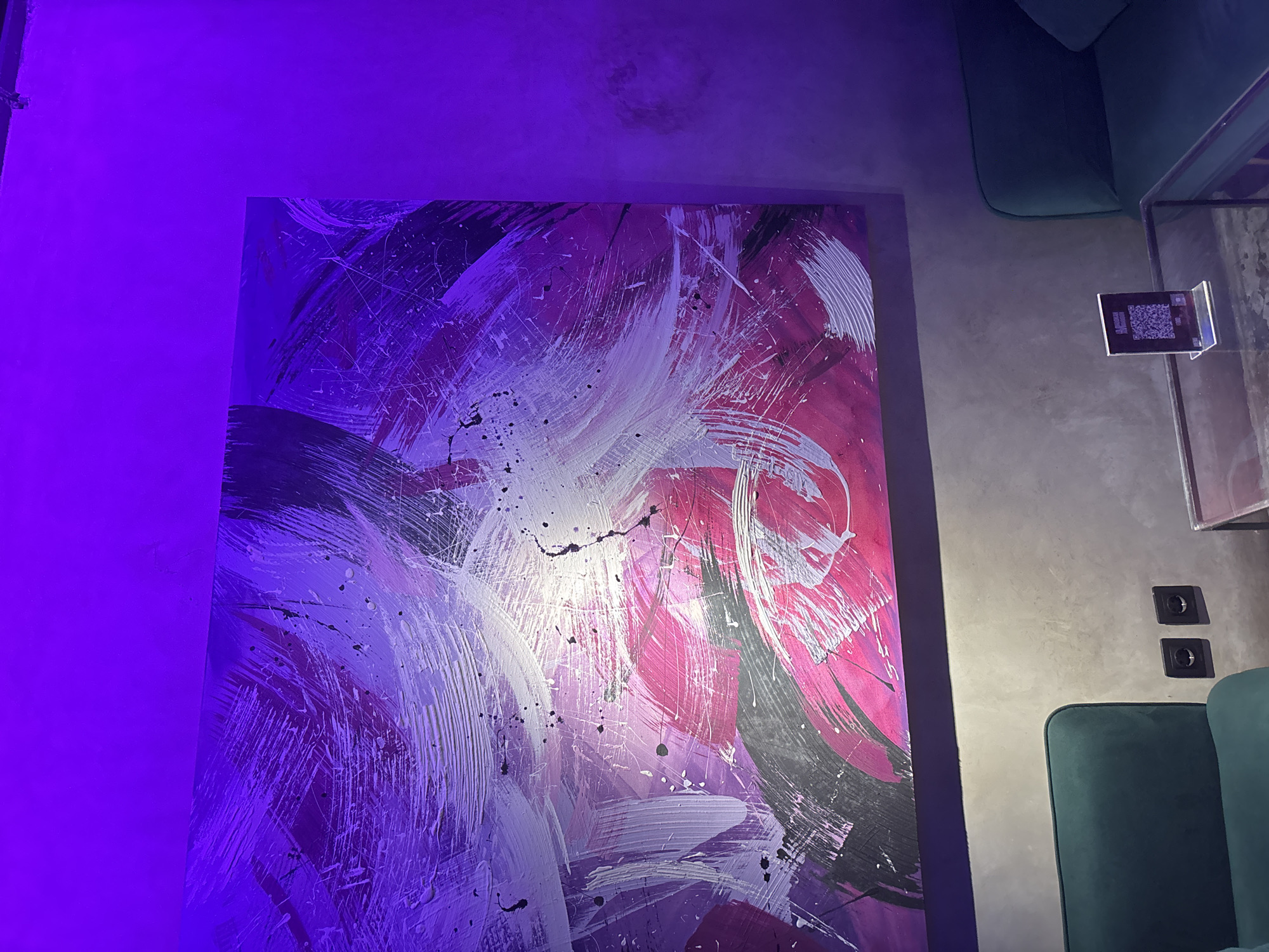 The image shows an abstract painting with broad strokes of purple, white, black, and pink. The painting is mounted on a textured gray wall. To the right of the painting is a teal-colored, upholstered armchair. A glass-topped table with a QR code display on it is visible next to the chair. Further down the wall are two electrical outlets. The location is Unknown, Indonesia. No people are visible.