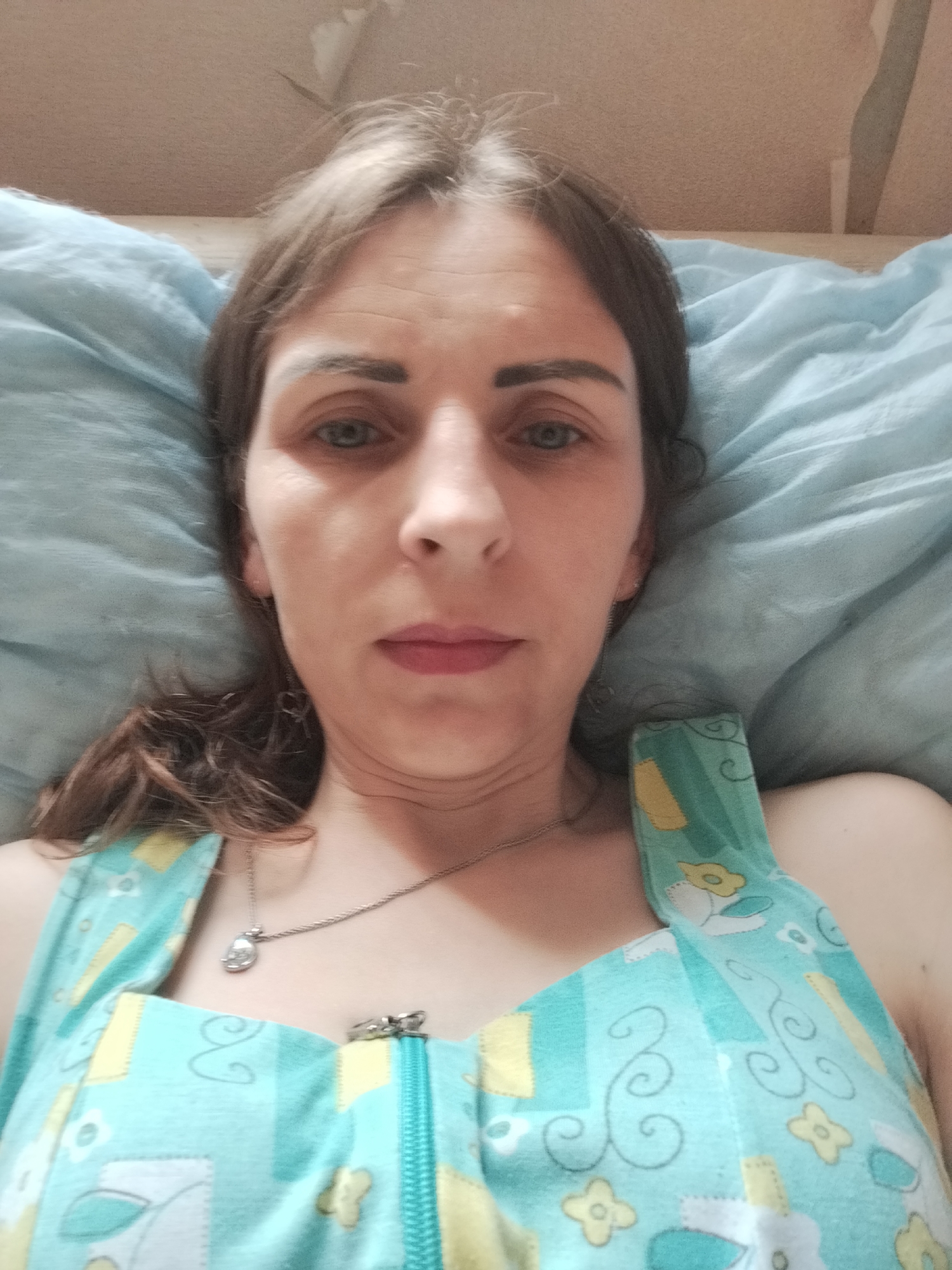 A woman lies on a light blue pillow, facing the camera. She has medium brown hair with some lighter streaks, and appears to be wearing a light blue and yellow patterned garment with a zipper. She is wearing a silver necklace with a small pendant. The background is a light brown surface, possibly a wall or ceiling. The scene is indoors. No location in Vinnytsia, Ukraine is identifiable.
