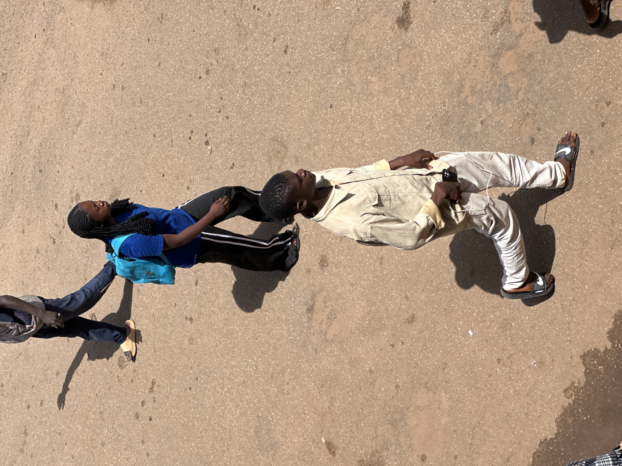 In Bukuru, Nigeria, three individuals walk on a dusty asphalt road under a bright sun, casting long shadows. In the background, a person in dark trousers and a light-colored top walks away from the viewer, wearing tan slides and a backpack. In the middle ground, a young woman with braided hair, wearing a blue t-shirt and black pants with white stripes, walks with a bright blue backpack. In the foreground, a young man in a light-colored button-down shirt and light-colored pants walks towards the right, holding a phone with white earbuds. He is wearing black Nike slides. The road surface is textured with small stones and shows evidence of water stains and scattered debris.