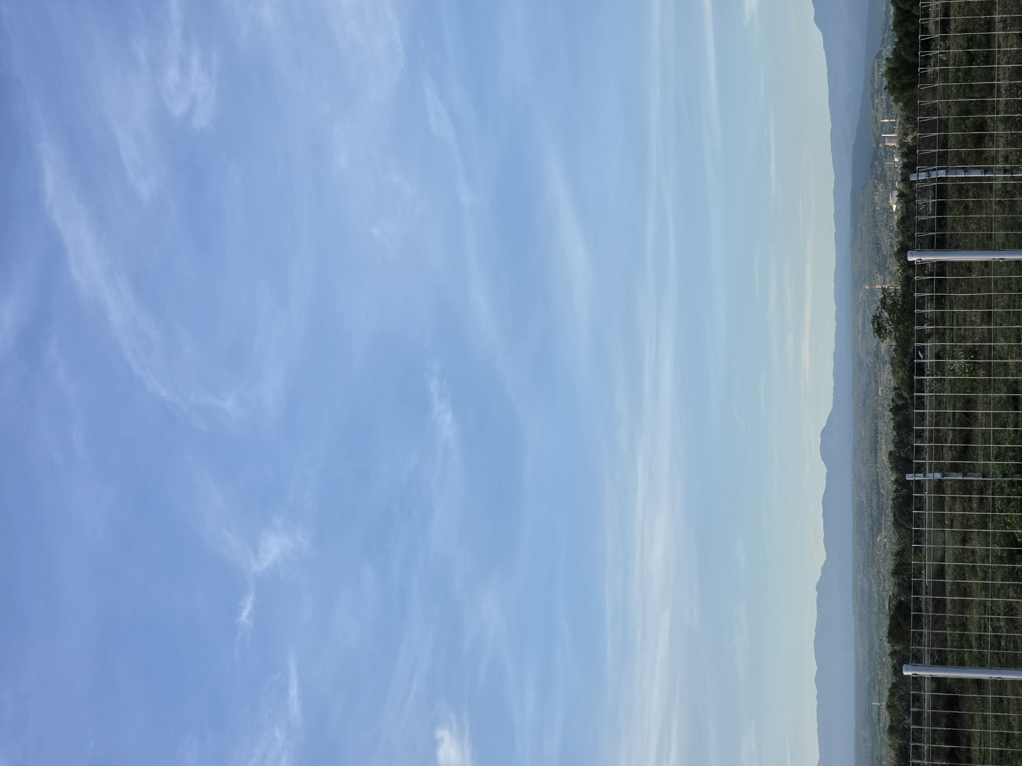 The image shows a view from Pintomakmur, Indonesia. A clear blue sky with wispy clouds occupies the left side of the frame. A mountain range is visible in the background, slightly obscured by haze. A wire fence runs along the right side of the image. A few buildings and vegetation are visible on the mountain side.