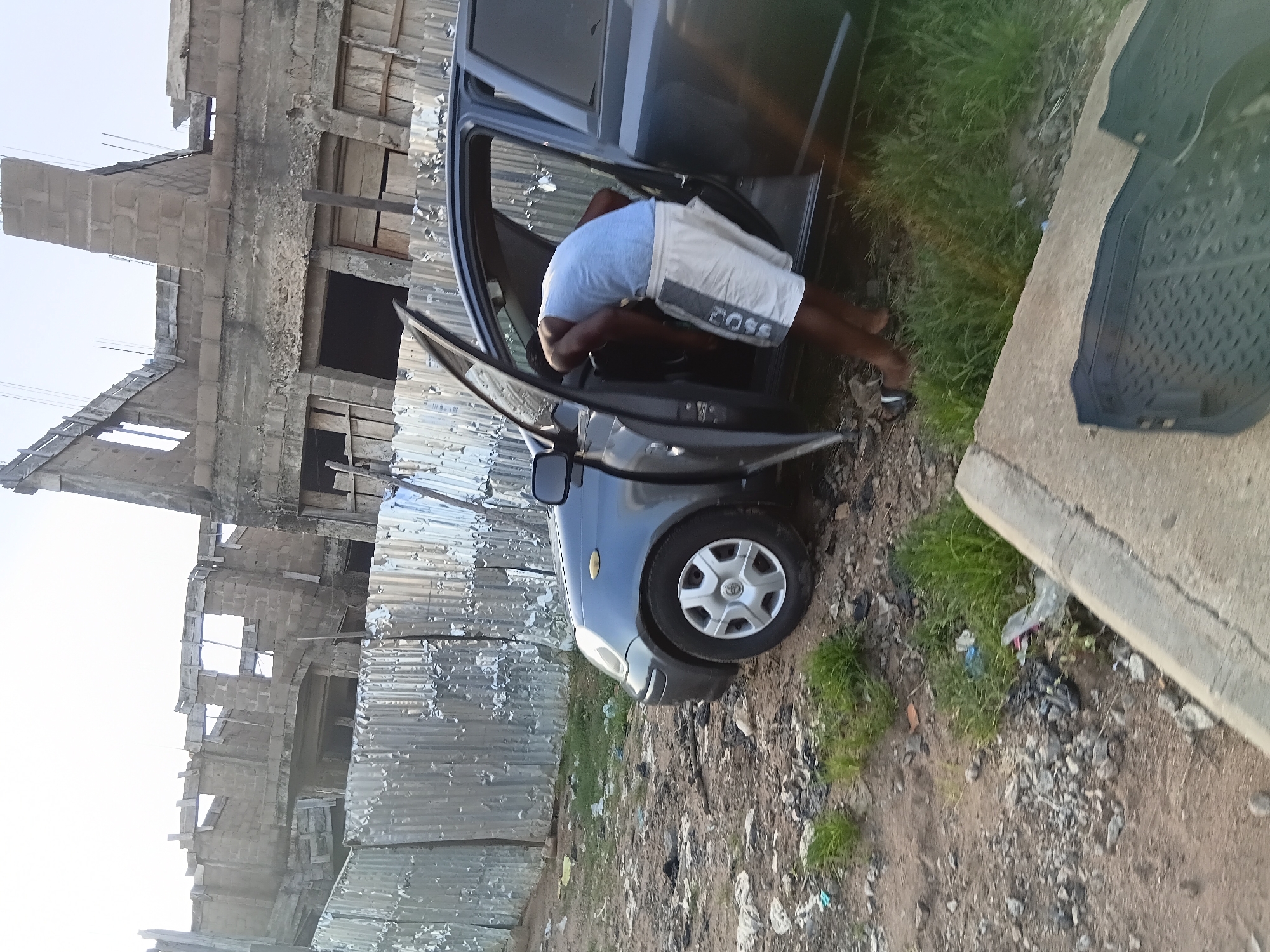 In Jalingo, Nigeria, a man in a light blue tank top and white shorts with a dark stripe and the word "BOSS" printed on it is leaning into the open driver's side of a dark grey hatchback car. The car is parked on a dirt and gravel lot with sparse green grass and debris. In the background, a corrugated metal fence separates the lot from an unfinished concrete building with exposed rebar and open window frames, suggesting construction is underway. The sky is overcast.