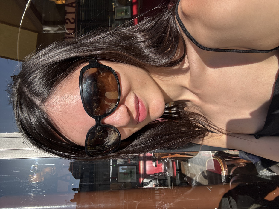 A female individual with long, dark hair and fair skin is depicted from the upper chest upward, positioned in front of a reflective glass surface. She wears large, dark-framed sunglasses with brown-tinted lenses, obscuring her eyes, and a black spaghetti-strap top. Her lips are slightly parted in a faint smile. Strong direct sunlight illuminates her right shoulder and face, casting defined shadows.
The reflective glass surface displays an urban outdoor environment and an indoor commercial interior. Reflections on the left show portions of distant light-colored buildings with windows, a clear blue sky, and a partial sign or text "CA". On the right, reversed text is clearly visible, reading "AT" and "ISTE", indicative of a "Brasserie" or similar establishment. The indoor reflections further reveal the interior of a cafe or restaurant, featuring red upholstered seating, dark wooden tables, and what appears to be part of a bar setup. The architectural style of the reflected buildings, combined with the presence of a "Brasserie" or similar French commercial establishment, places this scene in the city of Paris, France.