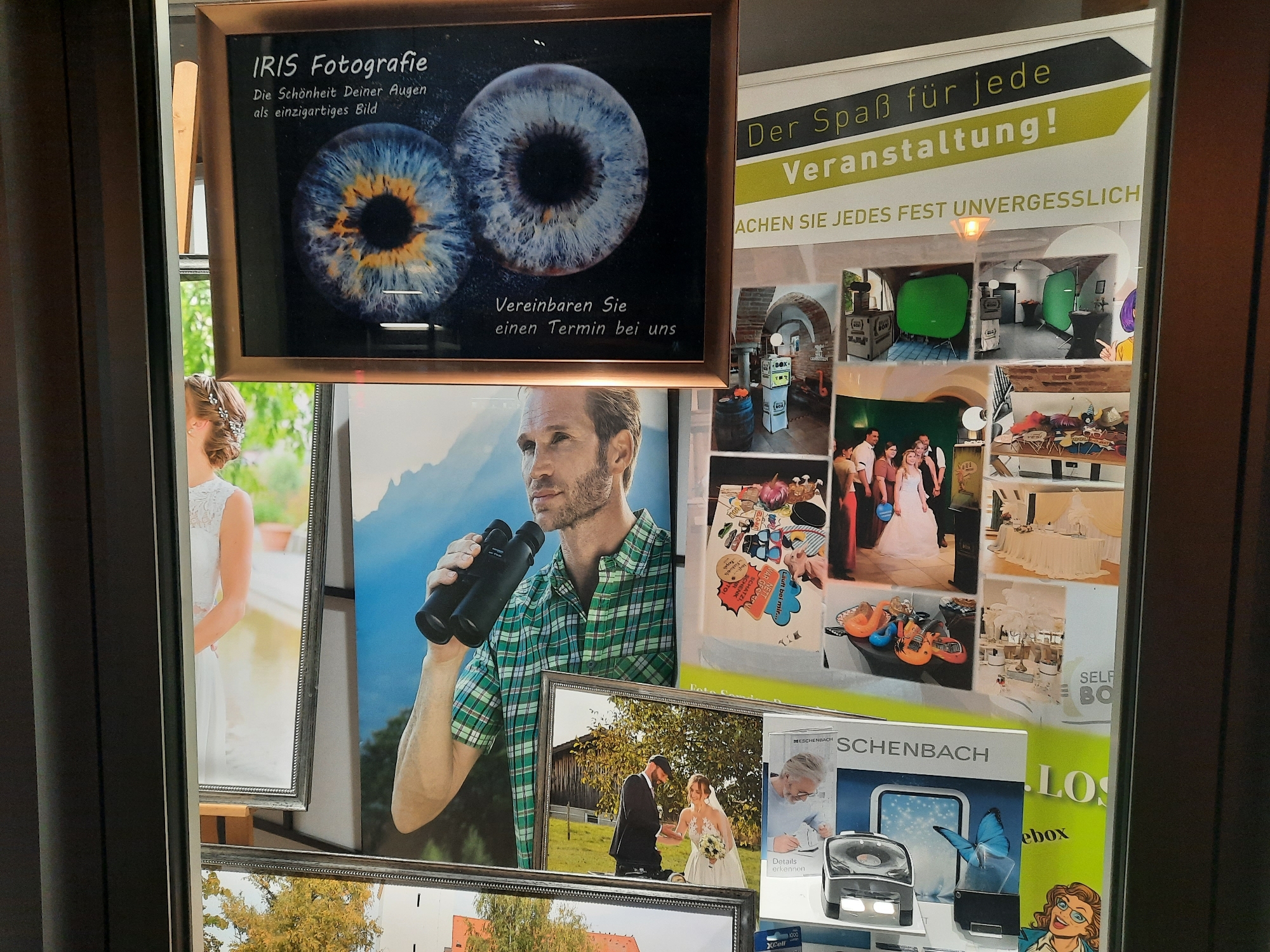 The image displays a window advertisement for "IRIS Fotografie" in Rosenheim, Germany. The advertisement features two close-up images of human irises with the text "Die Schönheit Deiner Augen als einzigartiges Bild" (The beauty of your eyes as a unique picture) and "Vereinbaren Sie einen Termin bei uns" (Make an appointment with us). To the right of this is a poster advertising "Der Spaß für jede Veranstaltung!" (Fun for every event!), with the tagline "MACHEN SIE JEDES FEST UNVERGESSLICH" (Make every celebration unforgettable). This poster is overlaid with smaller images depicting event setups, including a photo booth with a green screen, tables with decorations, and a group of people. Below the iris photograph is a framed image of a bride and groom. To the right of that, partially visible, is a framed image of a man holding binoculars. Further to the right, a display includes a product called "SCHENBACH LOS BOX" with images of a butterfly and a woman with glasses. The overall scene appears to be a commercial display, likely in a shop window, in Rosenheim, Germany.