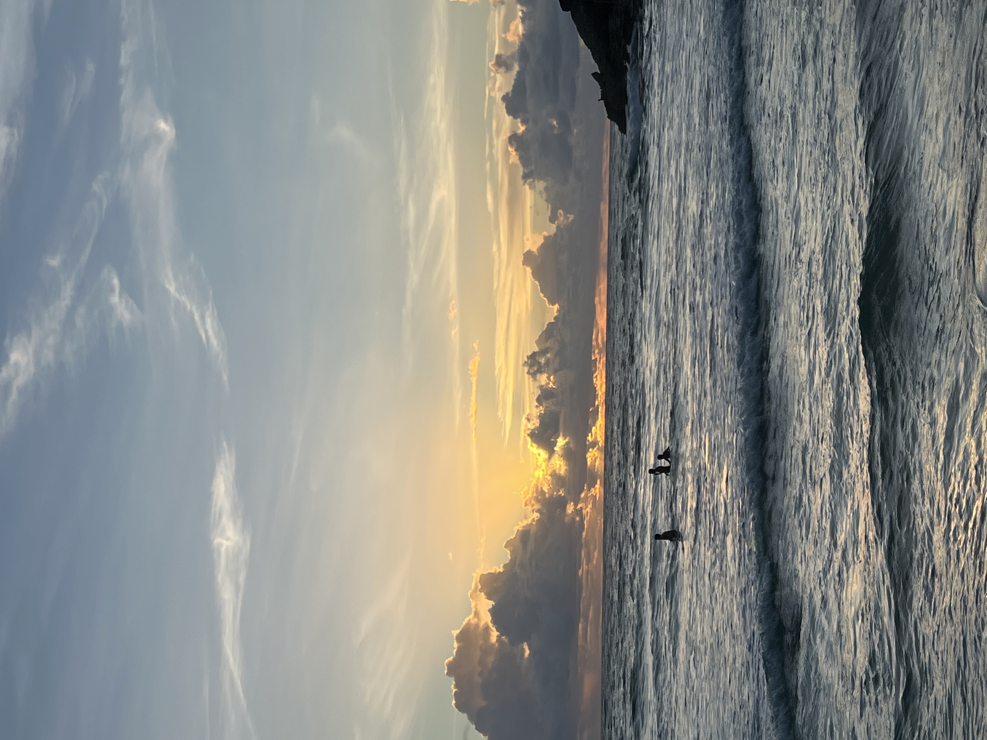 This is a landscape photograph taken from a low angle, depicting a beach scene at sunset. The image is oriented vertically.
The sky occupies the left half of the frame and is a soft blue, transitioning to a warm golden yellow near the horizon where the sun is setting. Wispy white clouds are scattered across the sky, catching the last rays of sunlight. The right half of the frame is dominated by the ocean. Gentle waves roll towards the shore, their surfaces reflecting the golden light of the sunset.
In the water, three silhouetted figures are visible. Two appear to be standing close together, while a third figure is slightly further away. They are likely people, possibly enjoying the sunset or wading in the water. The overall mood of the image is serene and peaceful, with the warm colors of the sunset creating a tranquil atmosphere.
The scene suggests a tropical location, possibly a beach in Thailand given the location context. The time of day is clearly sunset, indicated by the warm, low-angled sunlight and the illuminated clouds. There are no visible text elements in the image.