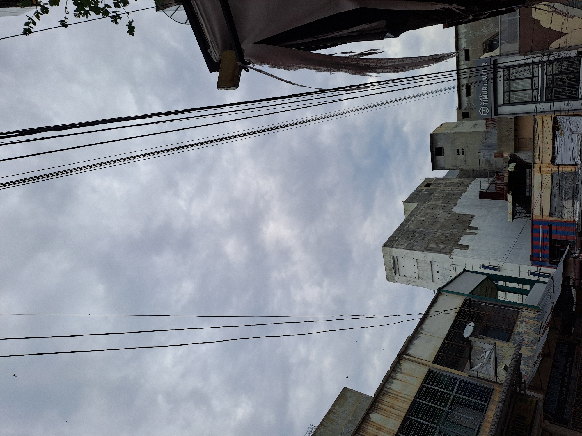 The image captures an overcast sky over a dense urban area, likely in Bandarbireuen, Indonesia, characterized by a clutter of overhead electrical wires and buildings. In the foreground and midground, several multi-story buildings are visible. The building on the far left has a weathered, aged facade with barred windows and a sign that appears to read "AQUEN PLASTIK." Adjacent to it is another building with a mix of white and grey concrete, showing signs of disrepair with patches of peeling paint and some windows. Further back, more structures rise, including one with a prominent sign for "APOTEK TIMUR LAUT 2." The overall impression is one of a bustling, possibly older, commercial district with a significant amount of exposed wiring contributing to a somewhat chaotic visual aesthetic. A few birds are present in the sky, and the general condition of the buildings suggests an environment that experiences significant weather exposure.