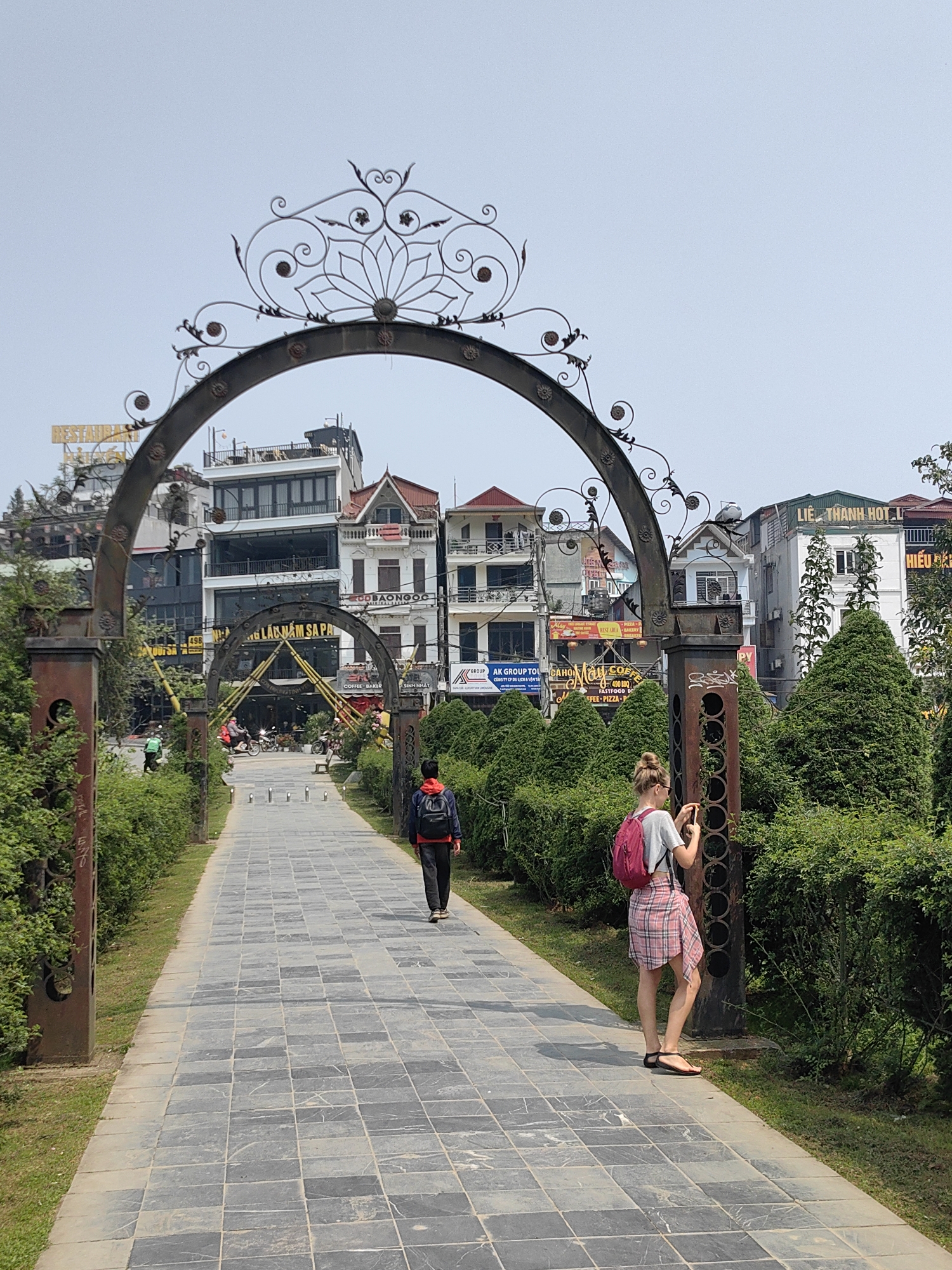 The image depicts a paved pedestrian pathway in Sa Pa, Vietnam, leading through a series of decorative metal arches towards an urban street. In the foreground, a fair-skinned female with blonde hair, wearing a gray t-shirt, pink plaid skirt, black sandals, and a pink backpack, stands to the right of the pathway. She is holding a smartphone with both hands, angled towards the right, appearing to photograph an unseen subject to her immediate right. She wears sunglasses.
Further down the pathway, slightly left of center, a person viewed from the back wears a red hooded jacket, dark pants, and a black backpack, walking away from the viewer. The pathway itself is composed of dark gray and lighter gray square tiles, bordered by green grass and manicured green bushes and hedges. Several ornate metal arches, exhibiting rust patina, span the pathway at intervals. The nearest arch features a central lotus-like design and decorative scrollwork.
In the background, beyond the pathway and subsequent arches, a dense urban scene unfolds with multi-story buildings. Visible signage on these buildings includes "RESTAURANT," "ĐẶC SẢN NÚI RỪNG S A PA" (Mountain Forest Specialties Sa Pa), "AK GROUP TOUR," "COFFEE - BAKERY," and "LIÊN THANH HOT. L." Architectural styles vary among the buildings, some featuring balconies and pitched roofs. Indistinct figures, including a person on a scooter/motorcycle and another individual, are visible on the distant street, indicating active traffic. The sky is bright and clear.
