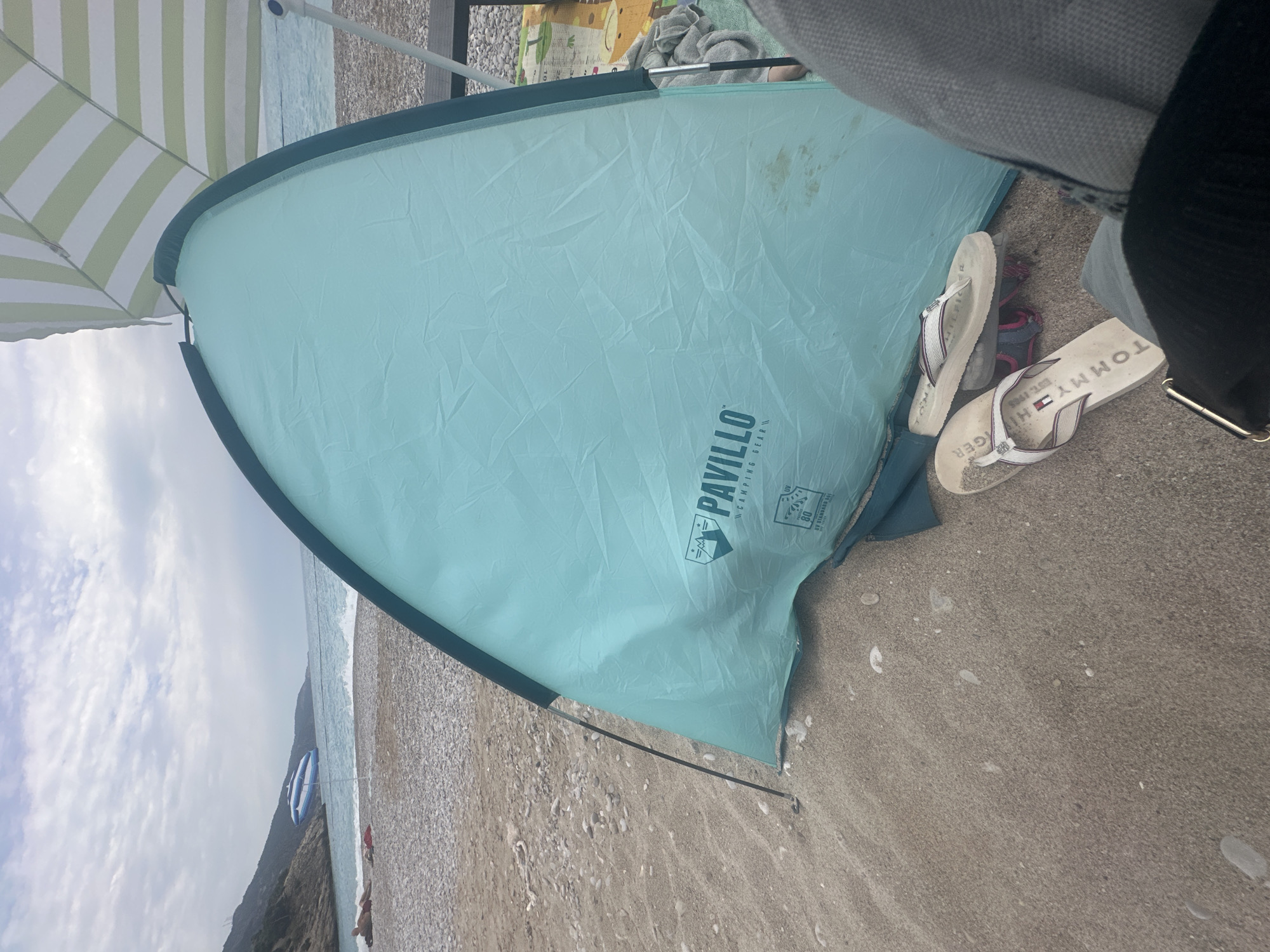 The image shows a beach scene in Fethiye, Turkey. A partially visible green and white striped parasol is to the left. A light-blue beach tent bearing the logo "Pavillo Camping Gear" sits on the sand. A pair of white Tommy Hilfiger flip-flops are placed next to the tent. The beach is made of dark sand with small shells and pebbles. The sea is visible in the background under a cloudy sky. A distant boat rests on the water. A coastal hill can be seen, with a beach chair at the water's edge, and an individual lying on the sand. The scene is somewhat obscured by a person's clothing on the right.