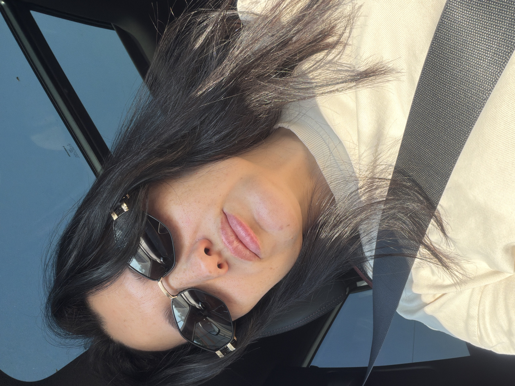 A woman with dark hair and sunglasses is sitting in a car, looking at the camera. She is wearing a light-colored shirt and a seatbelt. The car has a sunroof, through which the sky is visible. The overall lighting suggests it is daytime and sunny. The image does not contain any identifiable landmarks or clues that would allow for pinpointing a specific location within Noginsk, Russia, or any other city.