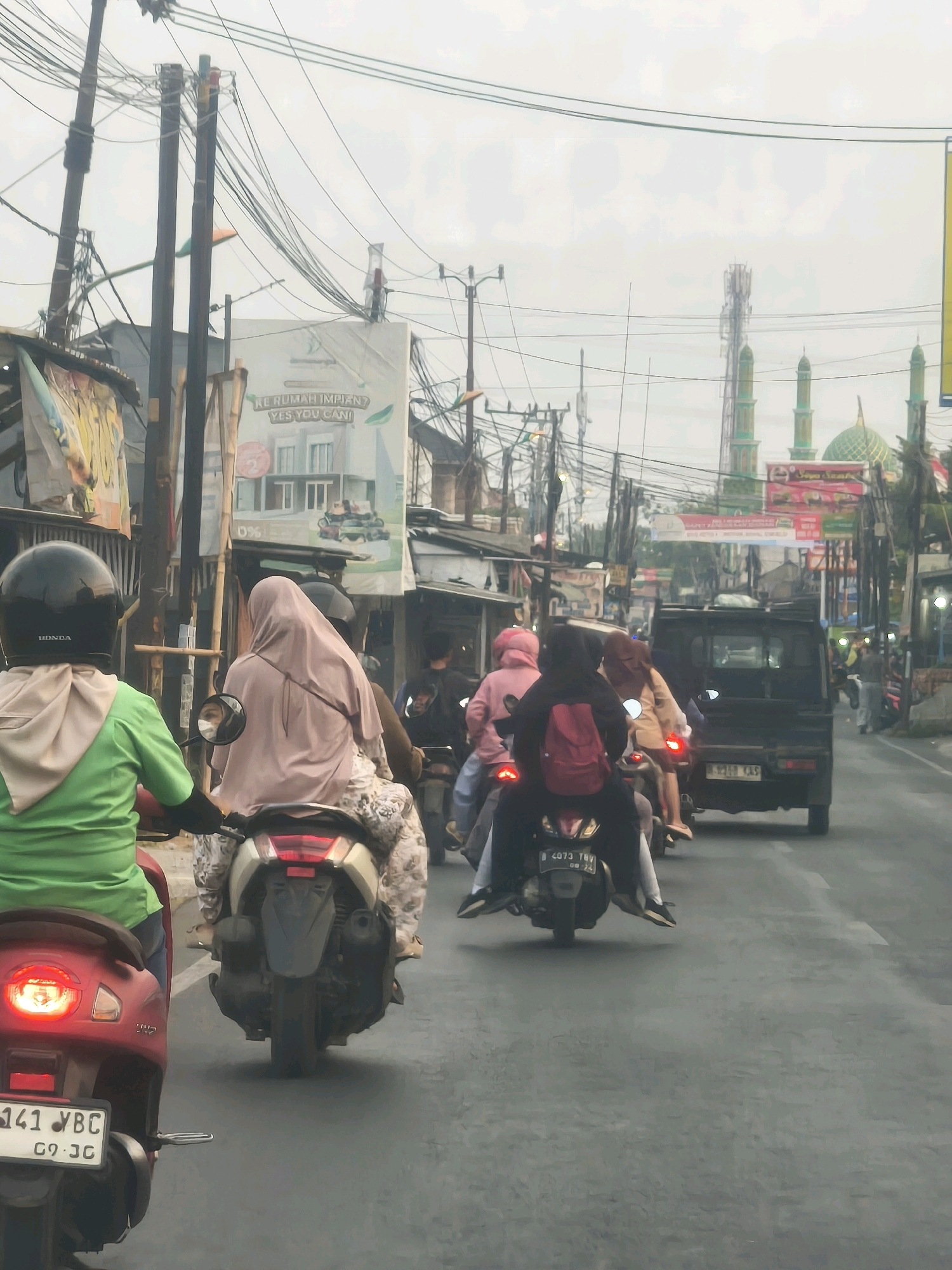In Satriamekar, Indonesia, a street scene unfolds with multiple motorcycles and a small truck navigating a paved road under an overcast sky. The foreground features a red motorcycle, with its brake light illuminated, and a person wearing a green shirt and a black helmet. Behind them, another motorcycle carries two individuals, one wearing a beige headscarf. Further down the road, a black truck is visible, followed by more motorcycles with passengers. The environment is characterized by a variety of buildings, utility poles with numerous wires, and advertising banners. Notably, a mosque with a green dome and minarets is visible in the distance, along with cellular towers. The overall impression is a busy urban street in Indonesia.