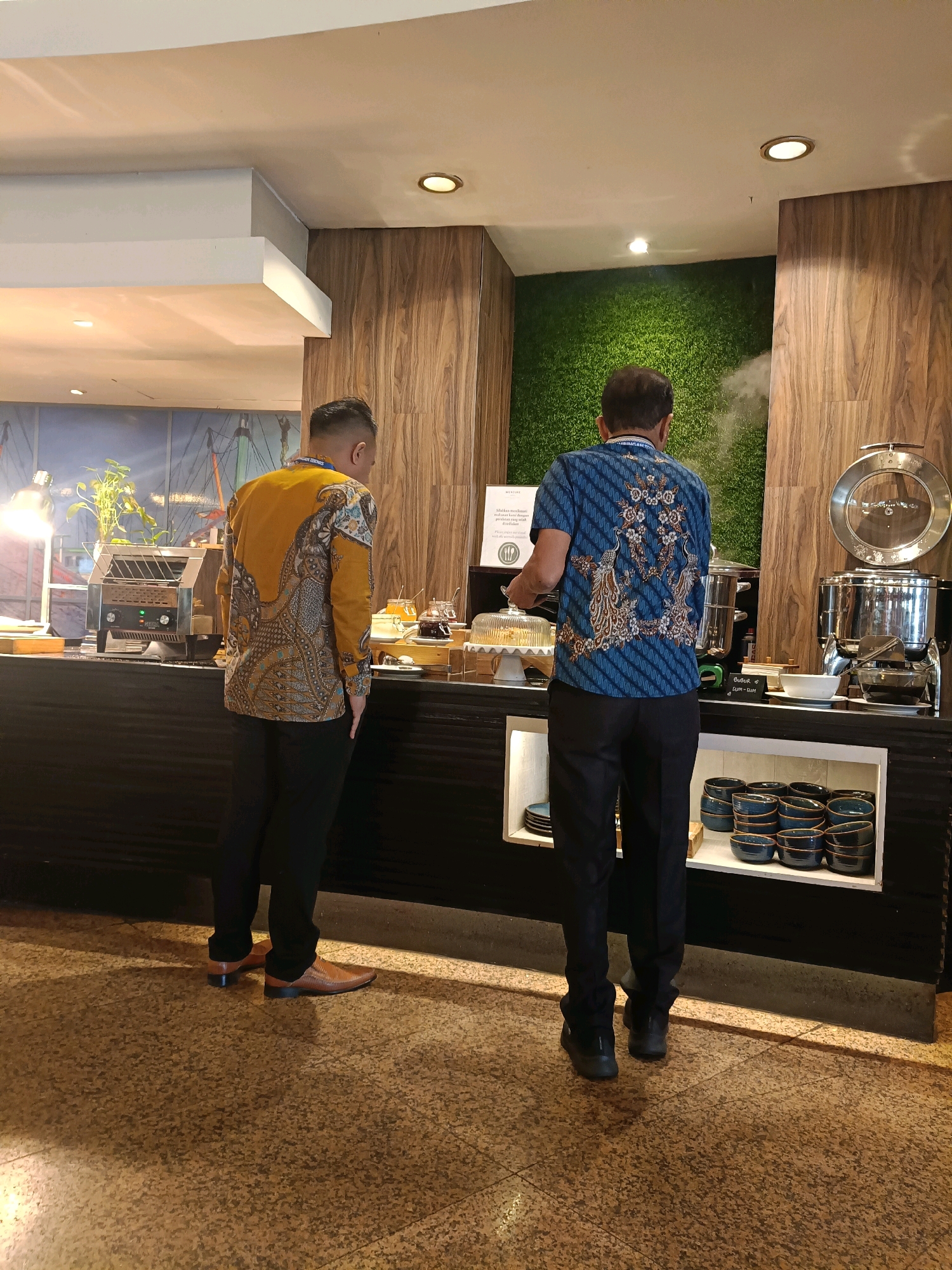 In North Jakarta, Indonesia, two men stand at a buffet. The man on the left, wearing a yellow batik shirt with intricate patterns and black pants, faces away from the camera, looking at a toaster. The man on the right, in a blue batik shirt with a peacock motif and black pants, faces the buffet, with steam rising behind him. The buffet features various food items, stacked bowls, and serving utensils, set against a backdrop of a green textured wall and wood paneling. The flooring is speckled brown.