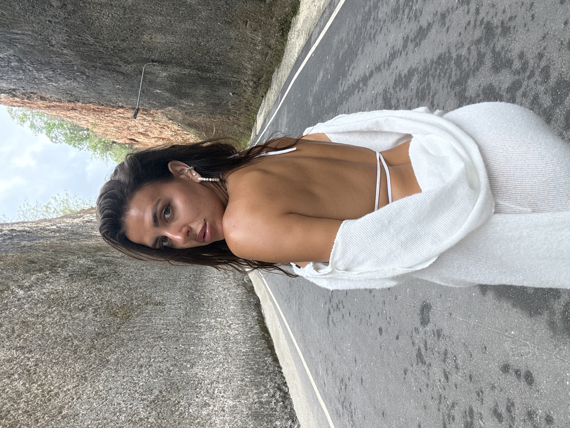 A medium shot captures a woman from behind, standing on a road with steep, rocky walls on either side. The road is paved and has a white line dividing it. The woman has dark hair and is wearing a white outfit. She is looking over her left shoulder at the camera. The rock walls are rough and textured, with some vegetation growing at the top. The sky is visible between the tops of the walls. The overall impression is one of natural beauty and a sense of isolation. This scene is likely located in the area of Kuta Selatan, Bali, Indonesia, known for its dramatic cliffside roads and beaches.