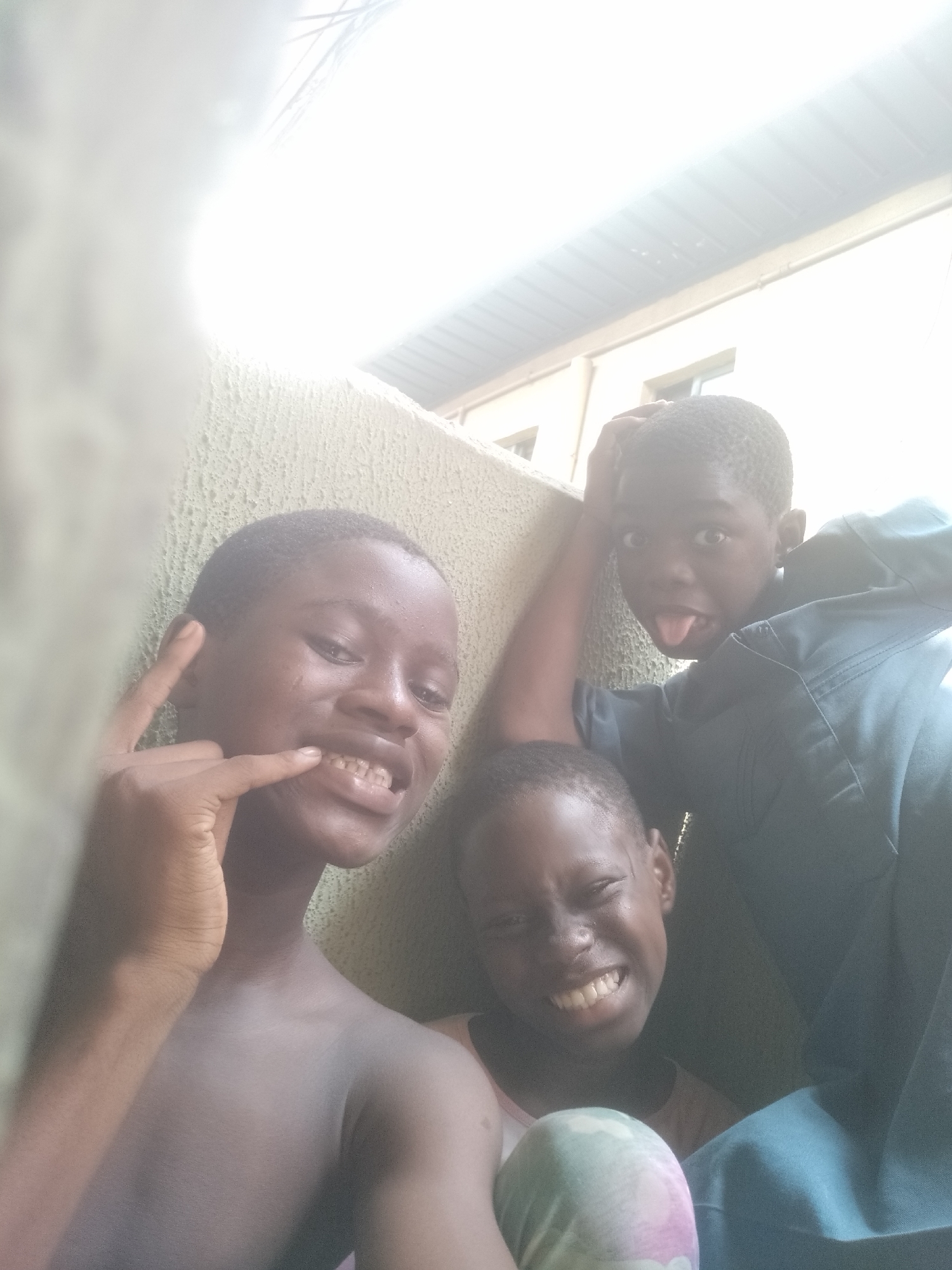 Three children, likely boys, are captured in a playful group photograph set against a textured, light-colored wall. The scene is brightly lit by natural sunlight, causing some overexposure in the upper part of the image, where hints of a building structure, including a window and roof, are visible. The setting appears to be an outdoor or semi-outdoor space in Shangisha, Nigeria, under a sunny sky.
On the left, one child, bare-chested, smiles broadly, revealing his teeth, and makes a hand gesture with his left hand, possibly a "rock-on" sign or pointing towards his mouth. In the center foreground, another child with short dark hair is grinning widely, eyes crinkled in joy, and appears to be wearing light-colored, patterned trousers. To the right, slightly behind the others, a third child wearing a blue top makes a humorous face by sticking out their tongue and holds one hand behind their head. All three children display expressions of happiness and playfulness. No visible text is present in the image.