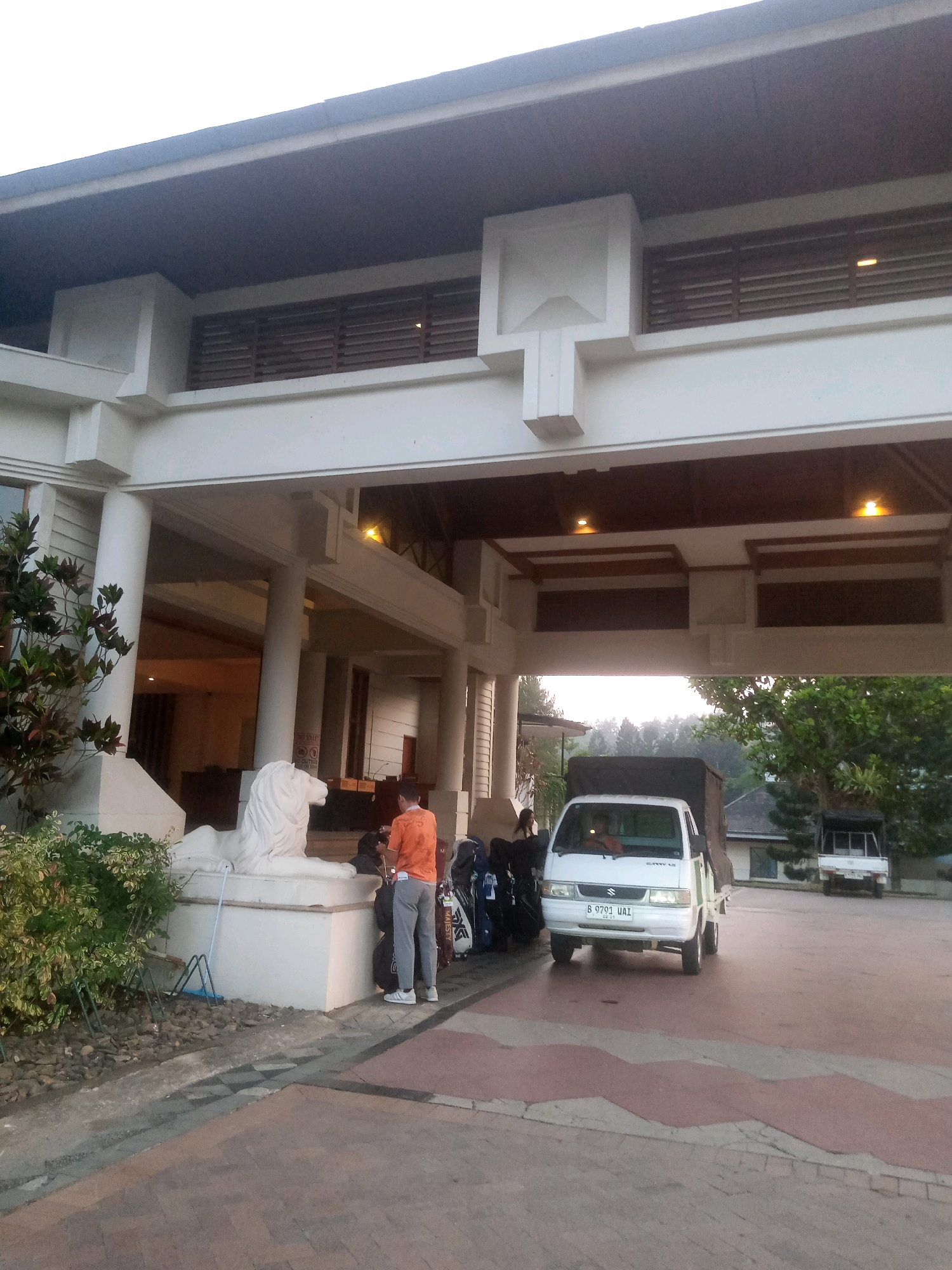 An exterior, low-angle shot captures the entrance of a resort or hotel in Babakan Madang, Indonesia. The building features a large overhang supported by thick white columns. Above, horizontal wooden slats are visible in recessed areas. A prominent white lion statue sits to the left of the entrance.
In the foreground, a white pickup truck with a canvas cover is parked under the overhang. A person wearing an orange t-shirt and grey pants stands near the truck, facing away from the camera. Several large bags, possibly golf bags, are stacked to the right of the lion statue and behind the man. To the left of the entrance, a person stands near a white truck parked further back. The ground is paved with interlocking bricks in a patterned design. The sky is overcast, and trees are visible in the background. The overall scene suggests a tranquil setting, possibly at dawn or dusk, with a sense of arrival or departure.