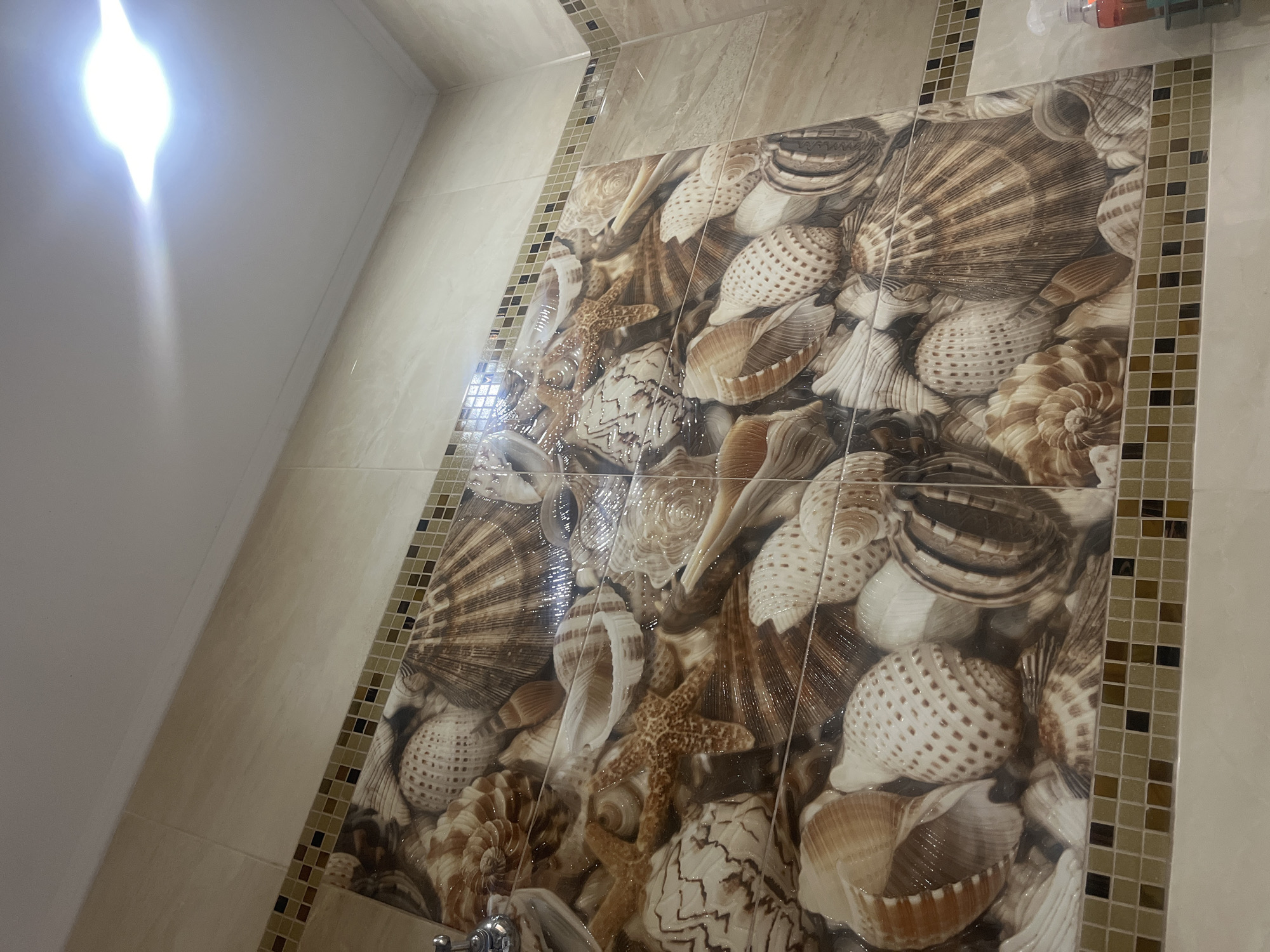This is a medium-angle, eye-level interior shot of a tiled wall, likely in a bathroom, featuring a detailed mosaic of seashells. The tiles themselves are a neutral, beige color, with a decorative border of small, square mosaic tiles in brown and beige hues just above the main seashell pattern. The seashell mosaic is printed onto the tiles and depicts a variety of shells and starfish in shades of brown, white, and cream, giving the impression of a beach scene. The lighting in the image appears to be from an overhead fixture, casting a bright, diffused light on the ceiling and reflecting off the glossy surface of the tiles. No people are visible in the frame. The overall impression is one of a decorative and perhaps themed bathroom wall. The location in Onokivtsi, Ukraine is not visually discernible from the image content.