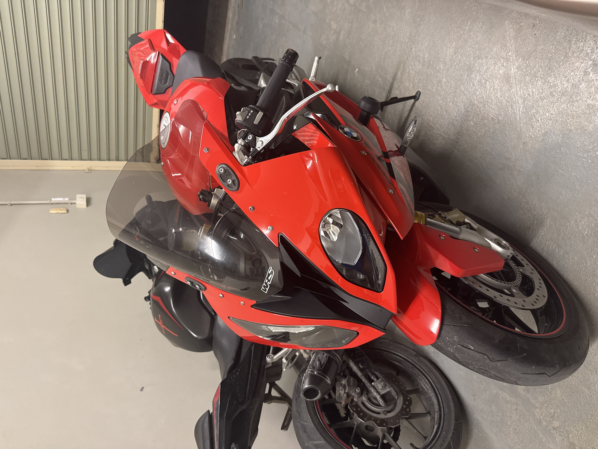 The image shows two sport motorcycles parked indoors in front of a closed metal roll-up door. The motorcycle in the foreground is bright red with black accents and a smoked windscreen. It features a visible BMW logo on the fairing and a carbon fiber exhaust on the rear wheel. The second motorcycle, partially obscured by the first, is black with red graphics and appears to be a sportbike as well. The environment is utilitarian, with a concrete floor and plain walls, suggesting a garage or storage area. The lighting is artificial and appears to be from overhead fixtures, casting soft shadows. No people are present in the image.