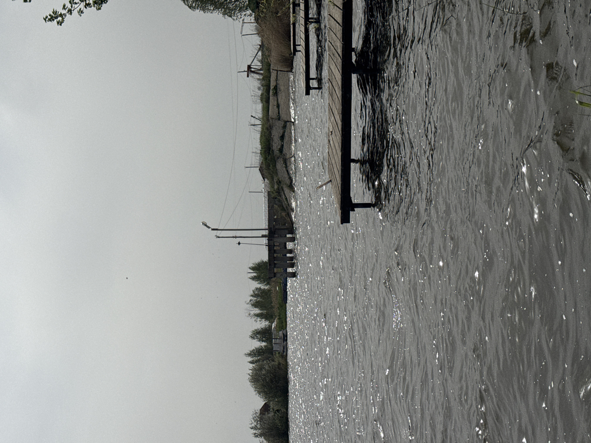 The image shows a body of water with a wooden pier extending into it. The water appears to be reflecting light, giving it a shimmering appearance. The pier is constructed of wooden planks and supported by metal posts. Along the shore, there is vegetation, including grass and trees. A light pole is visible further down the shore. The sky is overcast and gray. The location in the city is Unknown, Unknown.