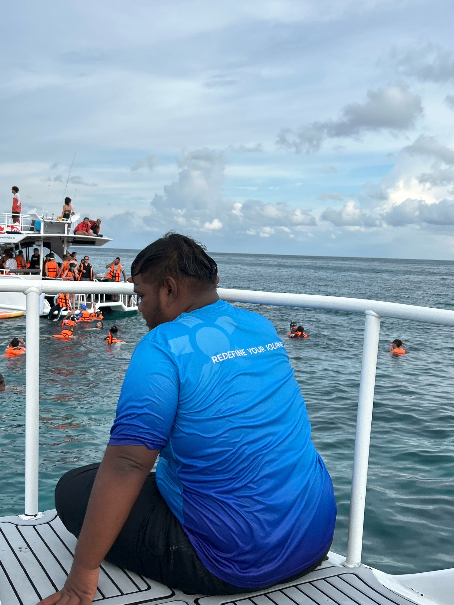 A person wearing a blue ombre t-shirt with the text "REDEFINE YOUR JOURNEY" printed on the back sits on the deck of a boat. In the background, a larger boat with several people is visible in the ocean, some wearing orange life vests. Other individuals, also in orange life vests, are in the water, appearing to be swimming or snorkeling. The sky is cloudy. The scene suggests a day trip or excursion in a coastal area. The location is identified as Rawai, Thailand.