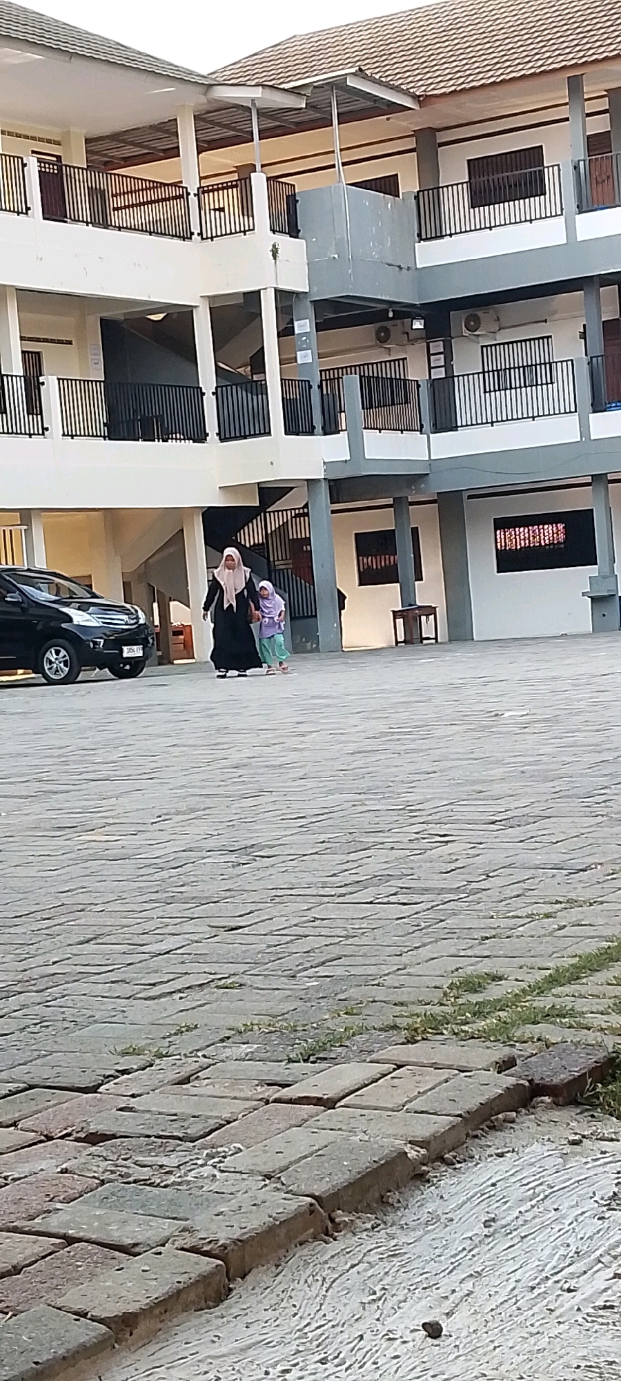 The image shows the exterior of a modern building with multiple stories, featuring white walls, gray accents, and a tiled roof. Black metal railings line the balconies and staircases. In the foreground, a paved courtyard with interlocking brick pavers is visible.
Two individuals are walking across the courtyard. The person on the left is an adult, wearing a black abaya and a light-colored hijab. They are holding the hand of a younger individual on the right, who is wearing a light purple top and light green pants, also with a head covering. A black car is parked to the left of the frame.
The environment appears to be an outdoor area of a school or institutional building. Based on the presence of air conditioning units on the building's exterior and the architectural style, it is consistent with a location in Karangrahayu, Indonesia. The lighting suggests it is daytime, possibly late afternoon given the shadows.