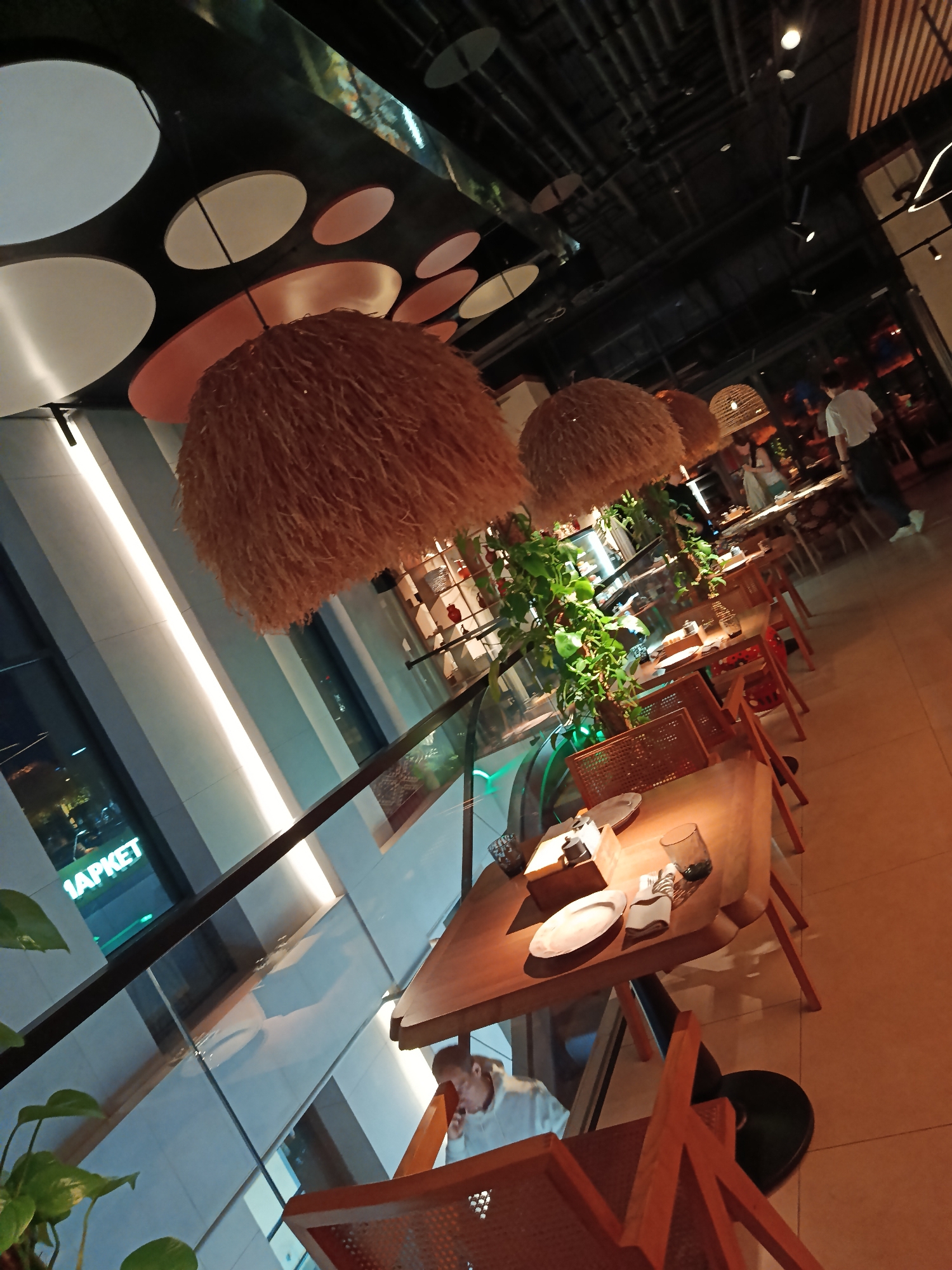 The image shows the interior of a restaurant located in Moscow, Russia, as indicated by the Cyrillic characters "1APKET" visible through the window. The restaurant features a modern design with wooden tables set for dining, each adorned with plates, glasses, and napkins. Seating consists of orange metal chairs. Above the tables hang distinctive straw-like lampshades. Green plants are interspersed along the tables, adding a touch of nature to the interior. A glass barrier separates the seating area from another level, where a person can be seen seated at a table, apparently on a phone call. In the background, other patrons and staff members can be observed. The ceiling is decorated with a pattern of large circles in white, black, and orange. The overall ambiance is modern and dimly lit.