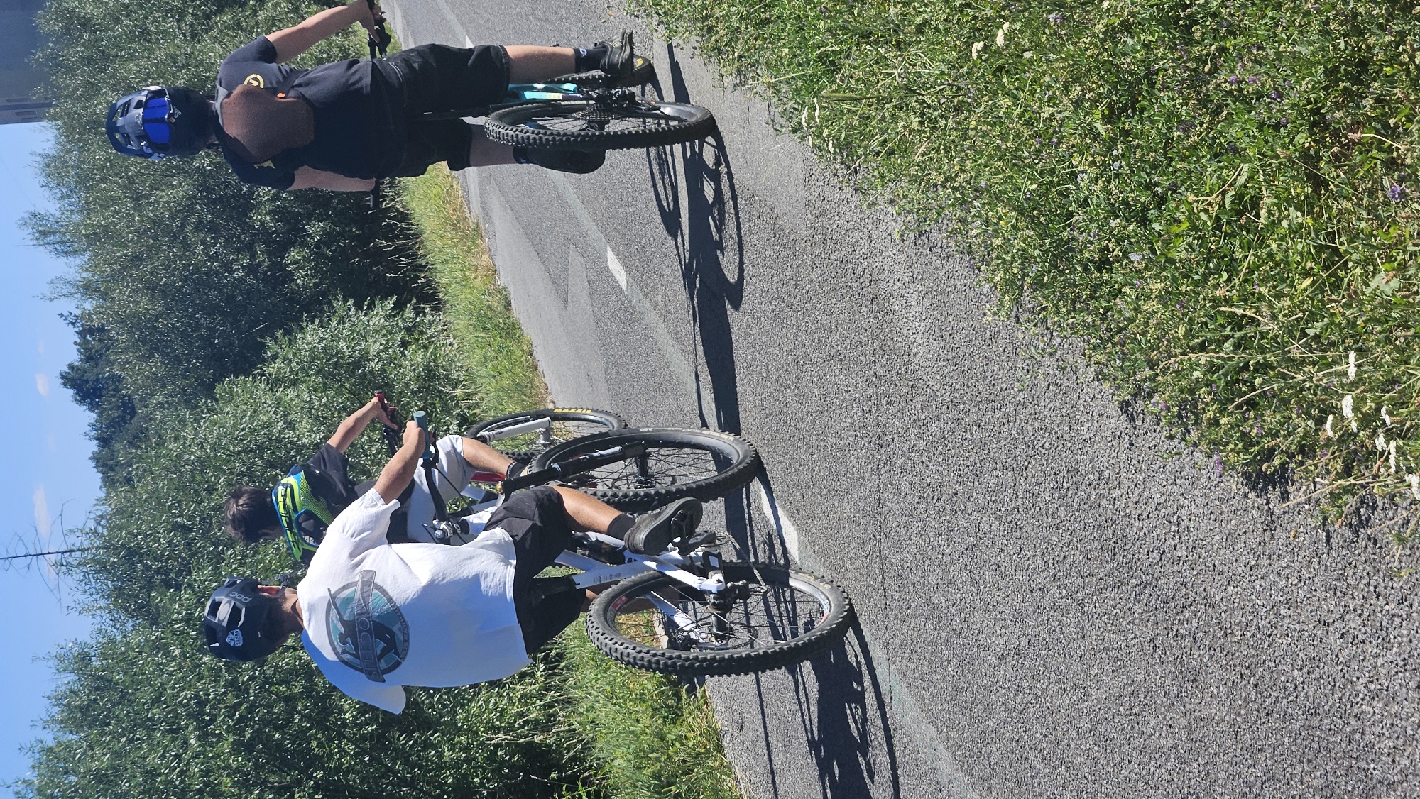 Three individuals are riding bicycles on a paved path adjacent to a wooded area under a clear blue sky. The person in the foreground is wearing a white t-shirt with a graphic and black shorts, and is riding a white and red bicycle. Behind them, another individual is wearing a black and neon green shirt and white shorts, riding a bicycle. The furthest individual is wearing a black t-shirt and black shorts, with a blue helmet and goggles, riding a blue bicycle. The path appears to be a dedicated cycling lane with white markings. The vegetation is dense green foliage. This scene could be consistent with a park or recreational trail within or near the city of Prešov, Slovakia, given the terrain and vegetation.