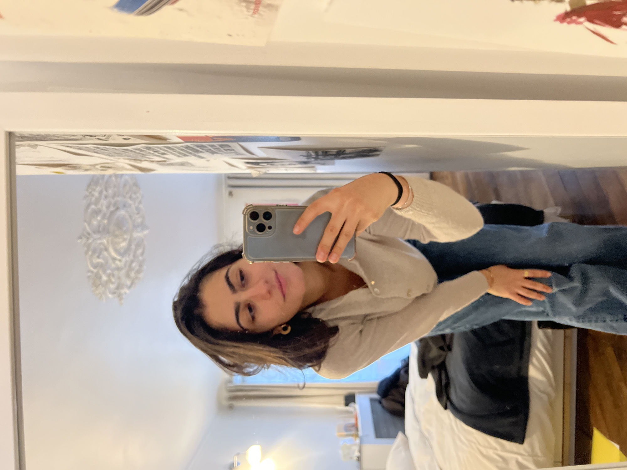 A woman is taking a mirror selfie in what appears to be an apartment in Paris, France. She is holding her smartphone in her right hand. She is wearing jeans, and a beige cardigan. A white decorative element can be seen on the wall behind her. A bed with white sheets is in the bottom right corner of the reflection. There is furniture visible further inside the room.