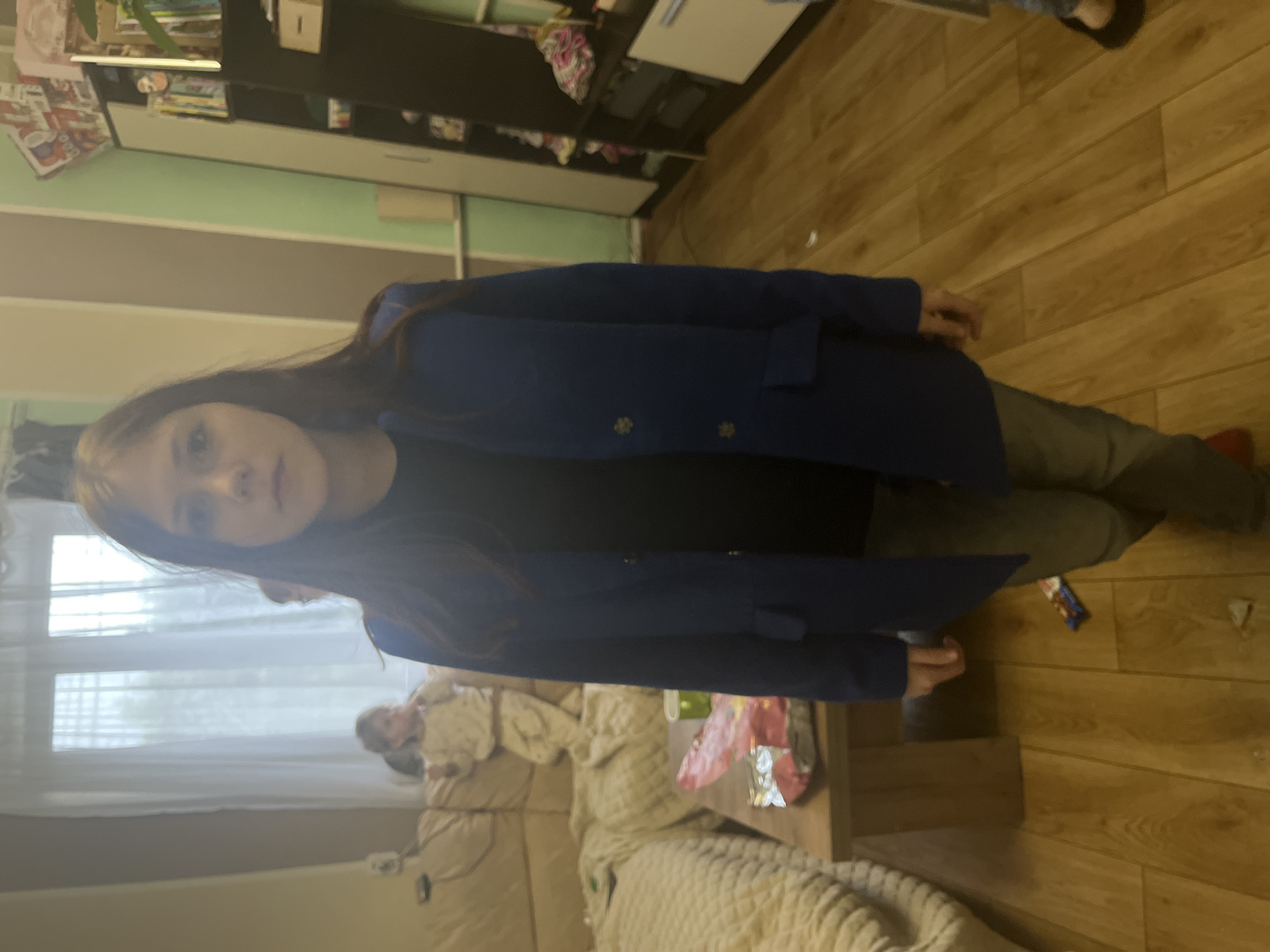 A child stands in a room with a window and a couch. The room appears to be in a home, with wooden floors and furniture. The child is wearing a blue coat. Another person is visible on the couch. The location is Oryol-Izumrud, Russia.