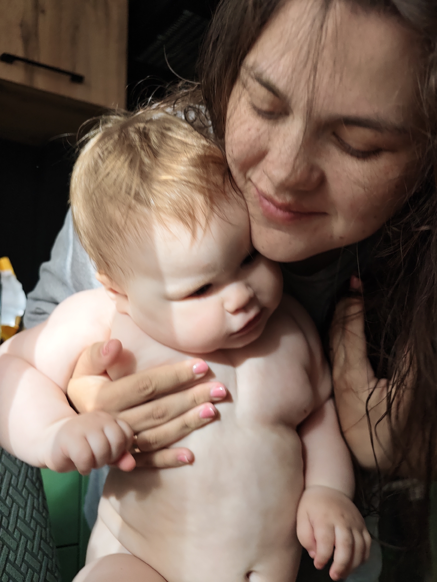 This intimate portrait captures a young woman holding a baby in what appears to be a home setting in Chaikovsky, Russia.
The primary subjects are a woman and a baby, depicted in a close embrace. The woman, positioned in the upper right, has long, dark hair, which appears somewhat disheveled. Her eyes are closed or looking down, and a gentle smile plays on her lips, conveying a sense of warmth, love, and contentment. Numerous freckles are visible across her nose and cheeks. Her left hand, adorned with light pink nail polish, is gently placed around the baby, and a subtle ring is visible on her ring finger.
The baby, with light blonde or light brown hair, is held in the woman's arms, facing away from the camera. The baby's bare upper body reveals soft, chubby features, characteristic of infancy. The baby's gaze is directed downwards and to the side. The woman's hand is supporting the baby, emphasizing the protective and nurturing connection between them.
In the background, elements of a domestic interior are visible. To the upper left, there's wooden furniture, possibly cabinets, with a dark handle. A small, light-colored bottle (possibly white or yellow) is partially visible behind the baby's head. A patterned fabric, possibly part of a chair or blanket, can be seen in the lower left. The overall lighting is soft and warm, contributing to the tender atmosphere.
No visible text is present in the image. The scene exudes an emotional tone of affection, care, and the deep bond between a mother and child.