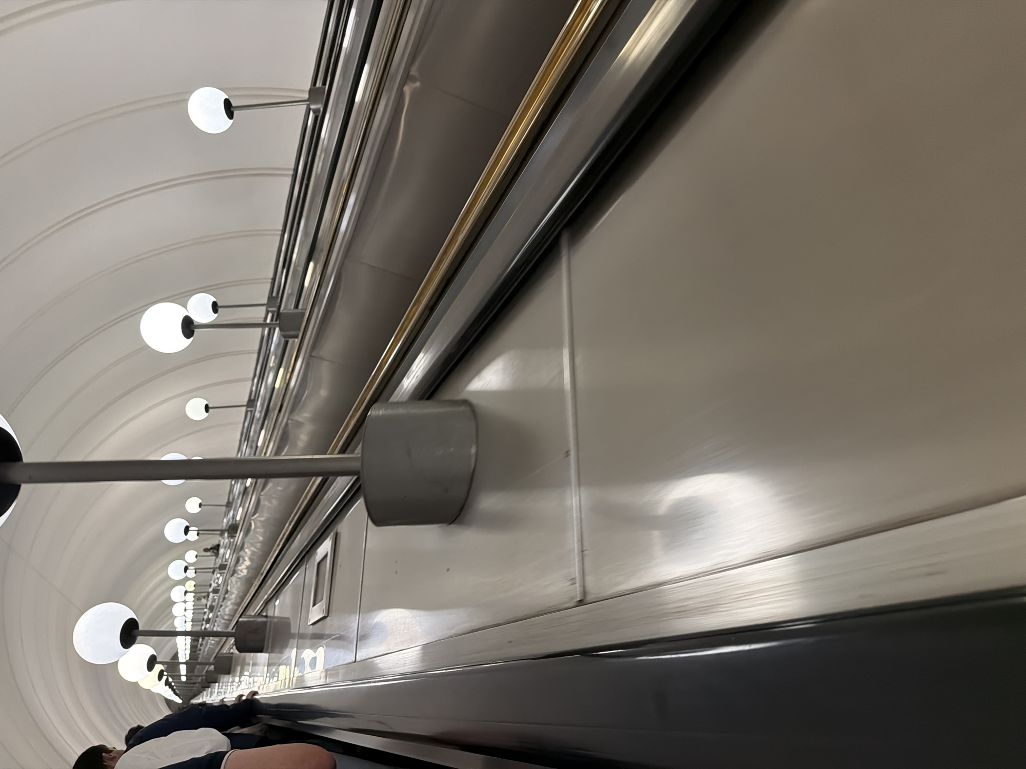The image shows a wide, long escalator in a subway station, characteristic of Moscow's Metro system. The escalator is stainless steel and illuminated by spherical white lights mounted on poles along its sides. The ceiling is a white, curved tunnel, also lined with lights. On the left side of the escalator, several people are visible, some standing and others moving upwards. The perspective is from a low angle, looking up the length of the escalator. The environment is clean and well-lit, suggesting an active public transportation hub.