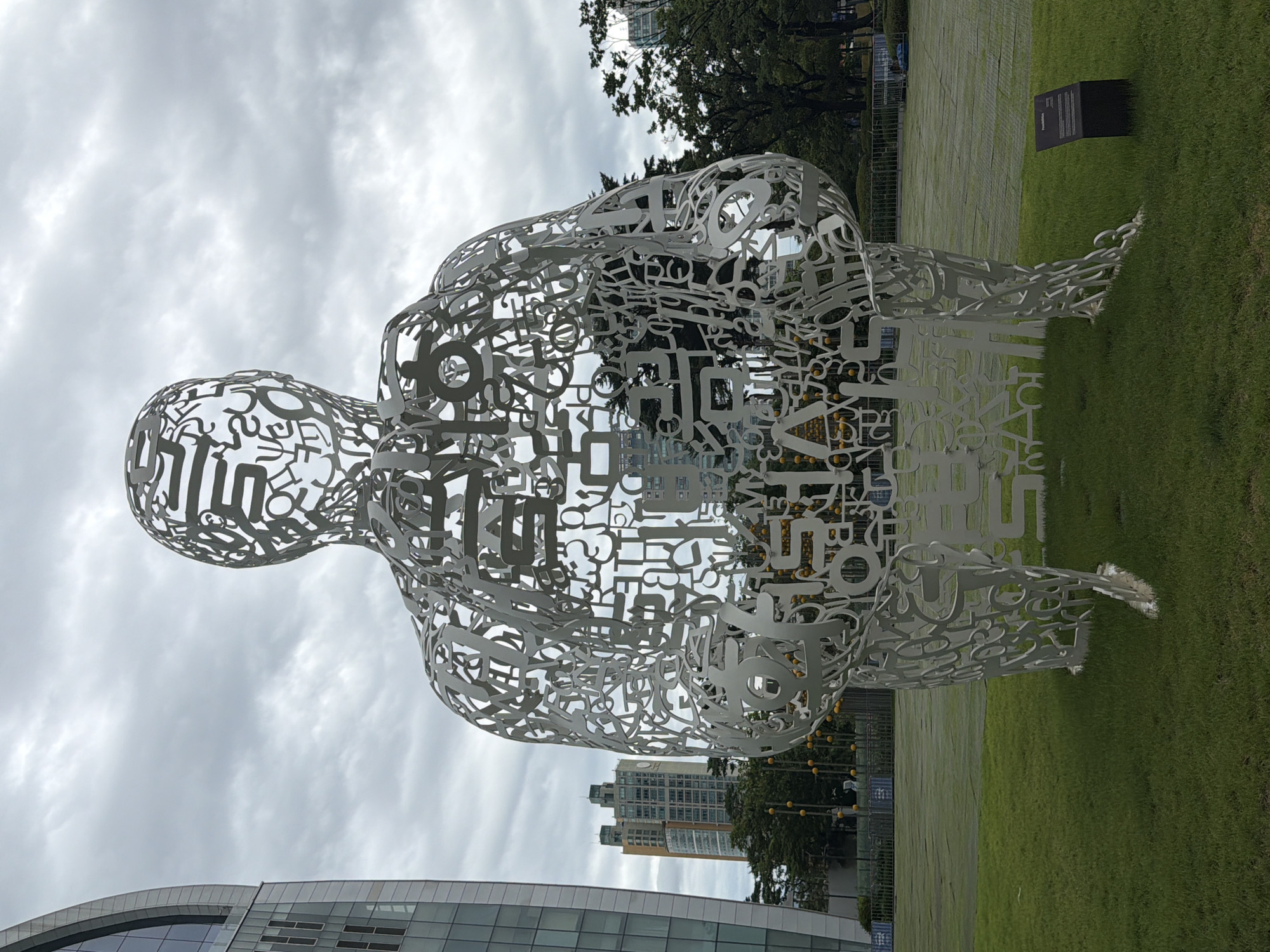 The image captures a large, open-air metal sculpture of a human figure composed of various letters and symbols, standing on a grassy lawn in Seoul, South Korea. The sculpture is white and intricate, with each part of the figure formed by a complex arrangement of stylized characters. Behind the sculpture, a modern building with a glass facade is visible on the left, and several taller buildings are seen in the distance on the right, suggesting an urban environment. Trees and a chain-link fence are also present in the background, separating the lawn from further structures. The sky is overcast with clouds. To the lower right of the sculpture, a small black informational plaque is mounted on the grass. The overall scene is outdoors, likely in a public park or plaza within the city.