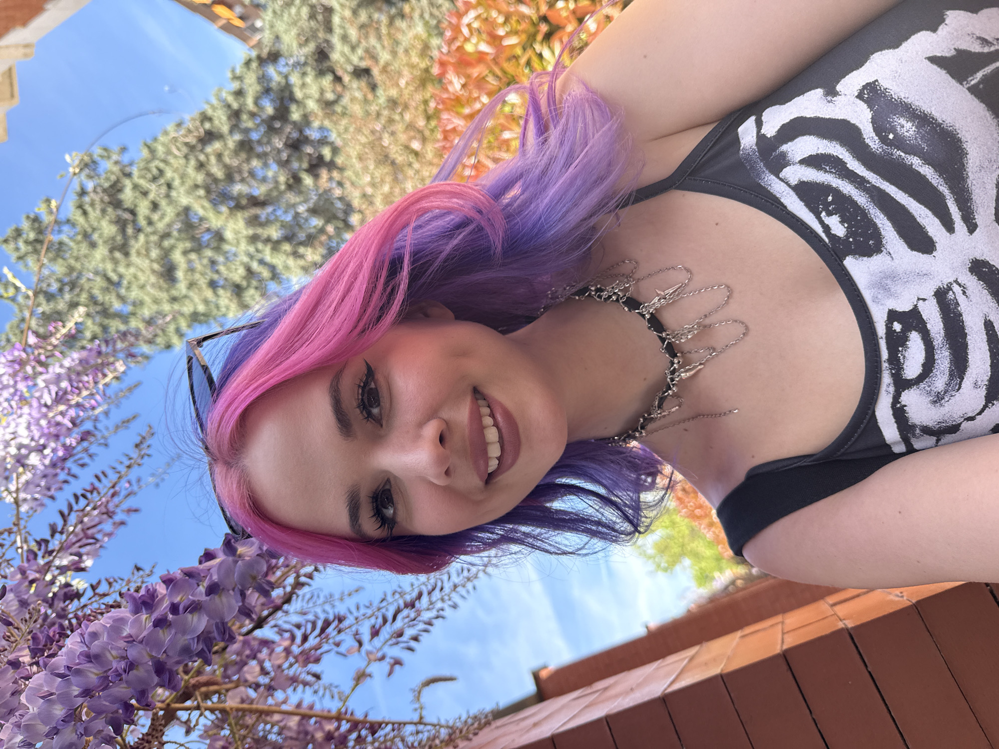 A woman with pink and purple hair smiles at the camera. She wears a black tank top with a white skeletal ribcage graphic and a chain necklace. Purple wisteria blossoms are visible in the lower left corner, and trees and the blue sky are in the background. The photo was taken in a place in Madrid, Spain, known for its wisteria flowers.