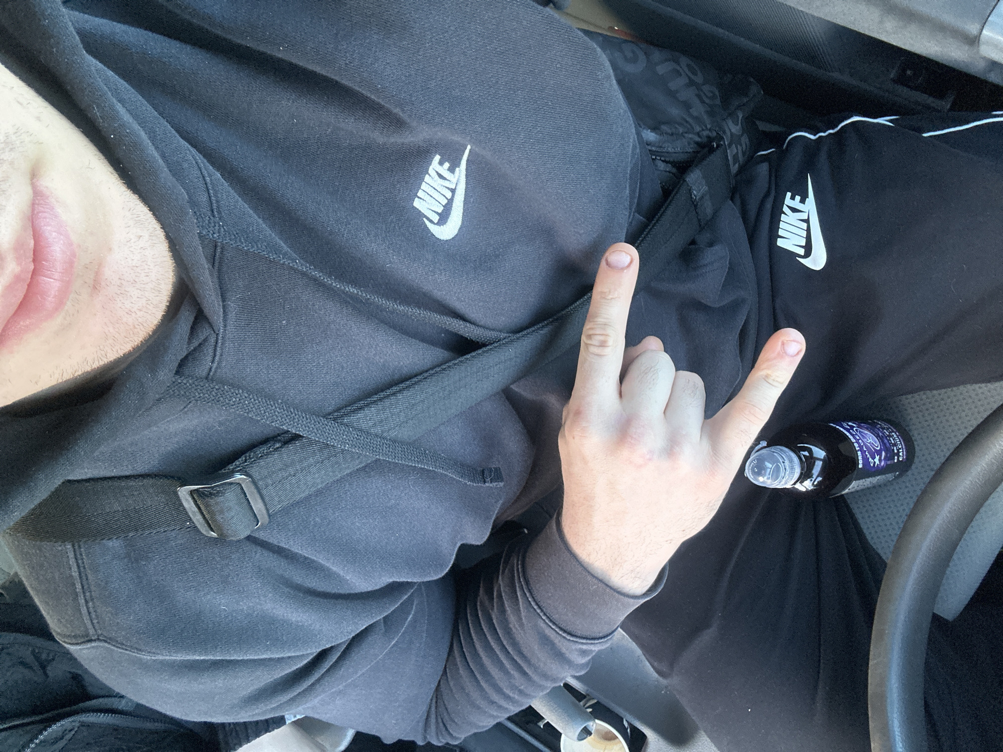 A photograph taken from a high-angle perspective shows a person seated, likely in a vehicle. The person is wearing a black Nike hoodie and matching Nike sweatpants, the brand logo visible on both garments. A black strap crosses the person's chest, suggesting a bag or carrying device. Their hand is raised, displaying the "rock on" hand gesture. A bottle of dark liquid rests on their lap, near the car's steering wheel. There is no information available from the image to indicate location in Burgas, Bulgaria.