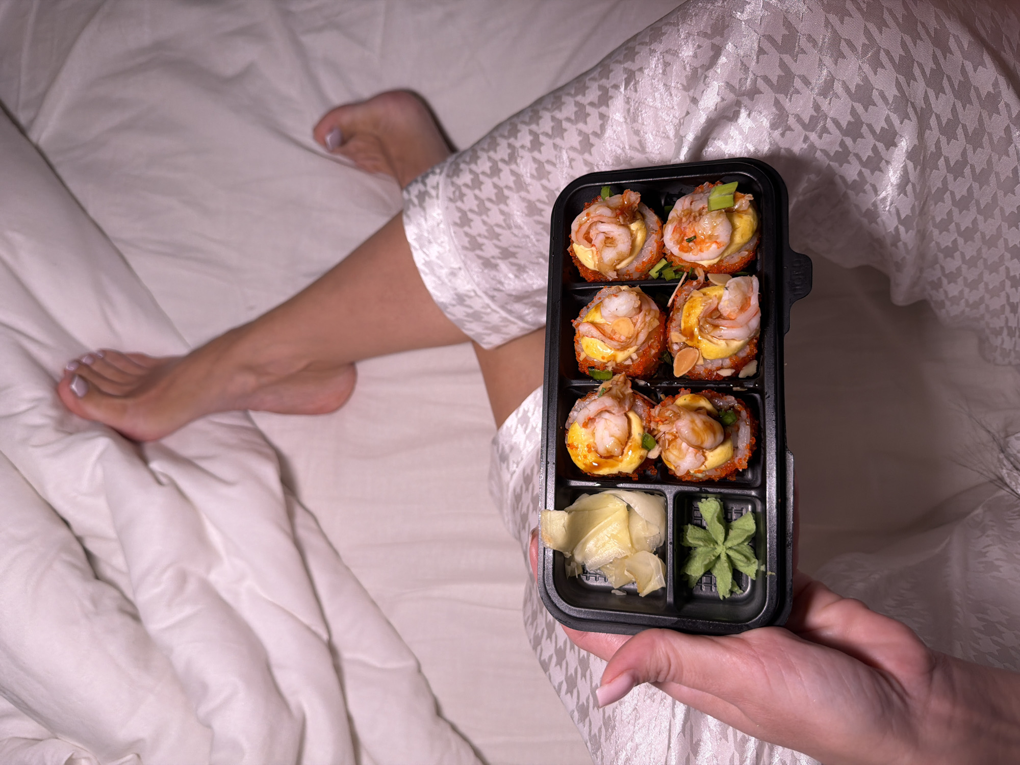 A person in houndstooth pajamas sits in a bed with white sheets, holding a black plastic sushi container. The container has six sushi rolls topped with shrimp and fish eggs, alongside a compartment with ginger and wasabi. The person's bare feet are visible in the periphery. There is no indication of the location being Svkh Voronezhskiy, Russia.