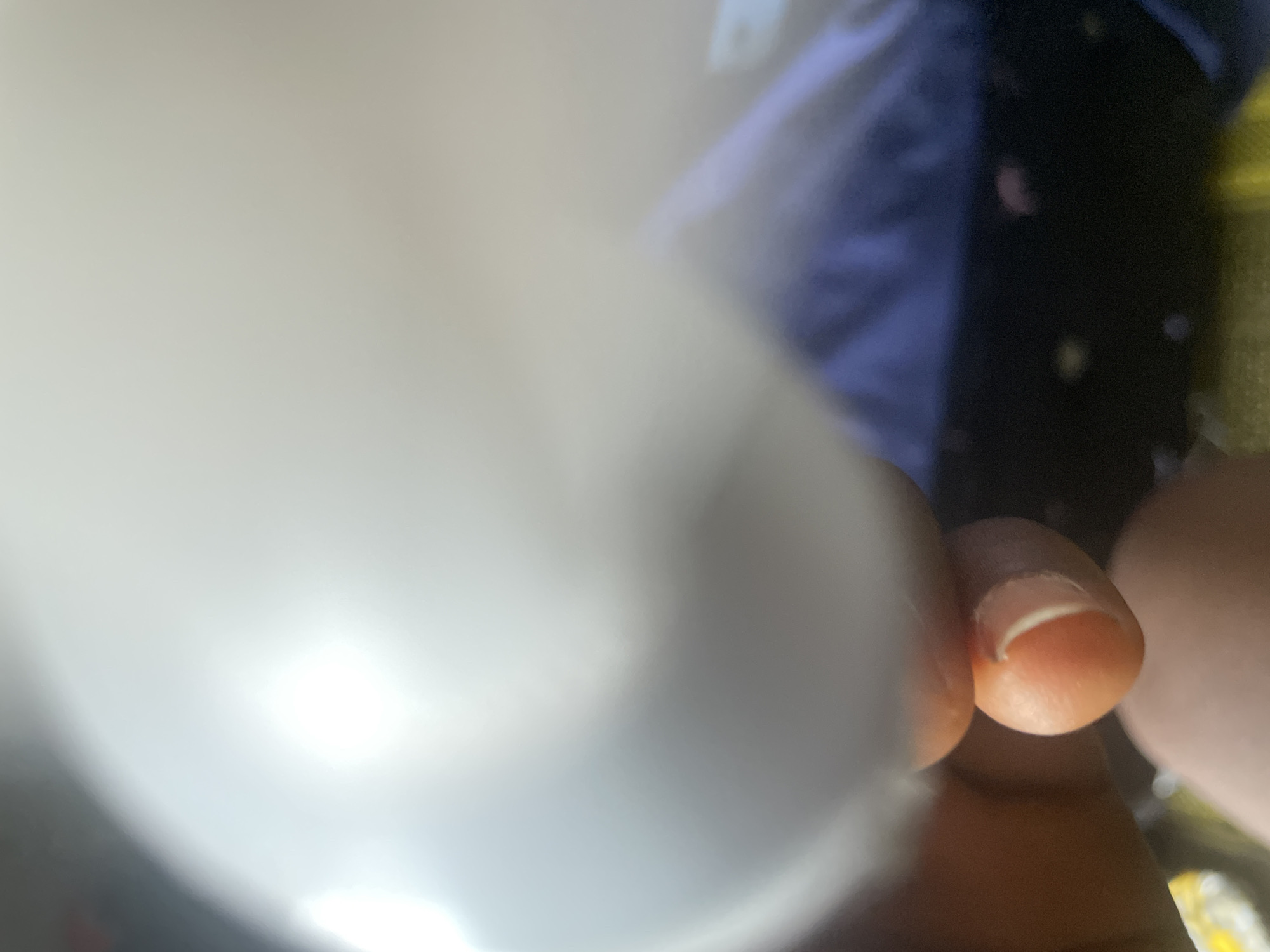 The image shows a close-up view of a person's fingers, with a focus on the fingernails of the index and middle fingers. The skin tone is medium to dark. The background is blurred, but appears to include a dark surface with some yellow and blue elements, possibly clothing or a bag. There is a large, out-of-focus white object dominating the left side of the frame, which could be a lamp, a part of a device, or even the camera itself. The lighting suggests it could be indoors, with a bright light source causing reflections on the white object and potentially on the skin. The overall impression is intimate and personal, focusing on the details of the hand. No text is visible in the image. The context of Jalingo, Nigeria, does not provide specific visual cues within the image itself.
