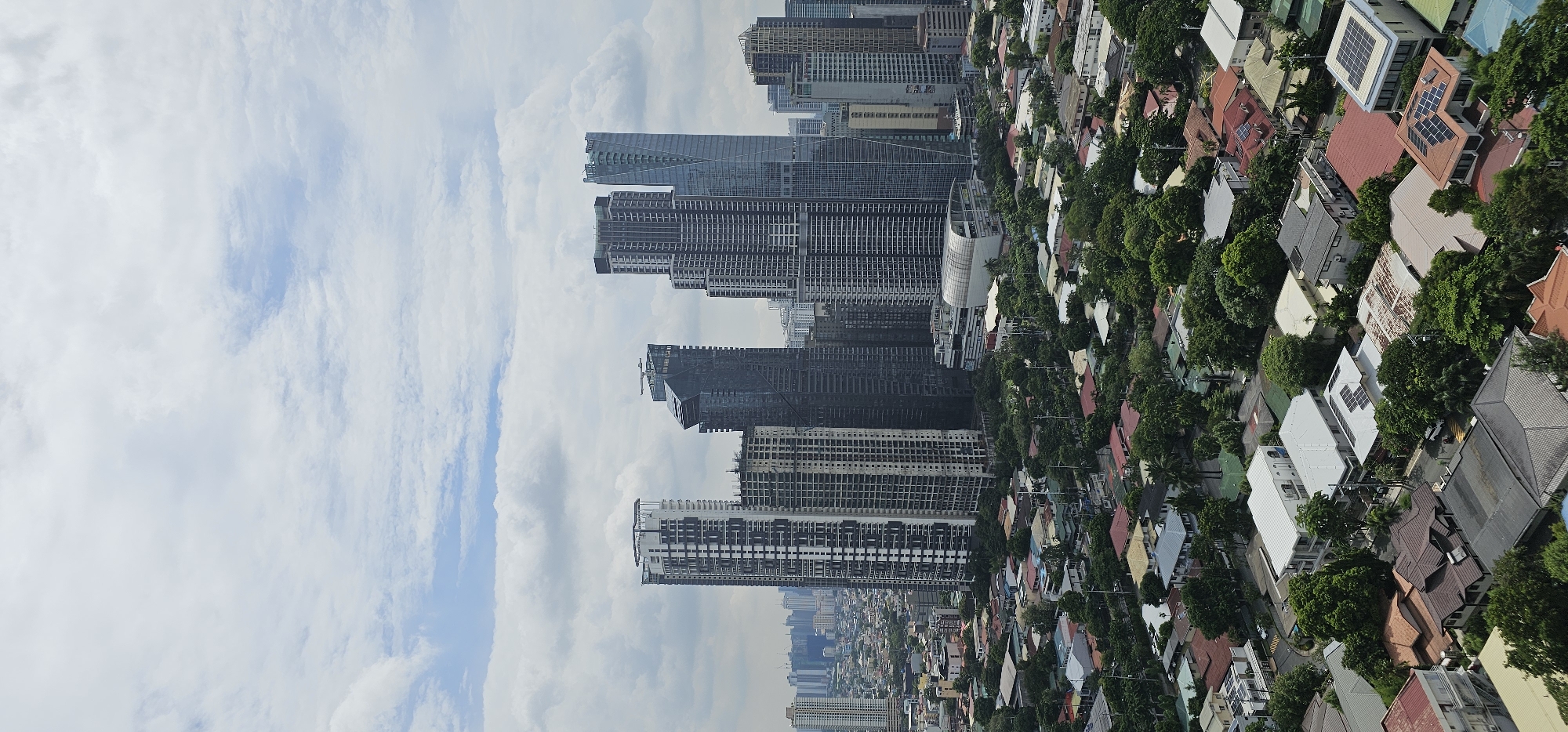 This is an aerial, vertical, medium shot of the cityscape of Makati, Philippines, on a cloudy day. In the background, several high-rise buildings dominate the skyline, with varying architectural designs and heights, indicating a commercial district. In the foreground, a dense residential area with numerous houses featuring red and grey tiled roofs, interspersed with lush green trees, stretches across the landscape. Some of the houses are equipped with solar panels. The image captures a contrast between the modern urban development of high-rises and the more traditional housing in the residential areas. There are no people visible in the image.