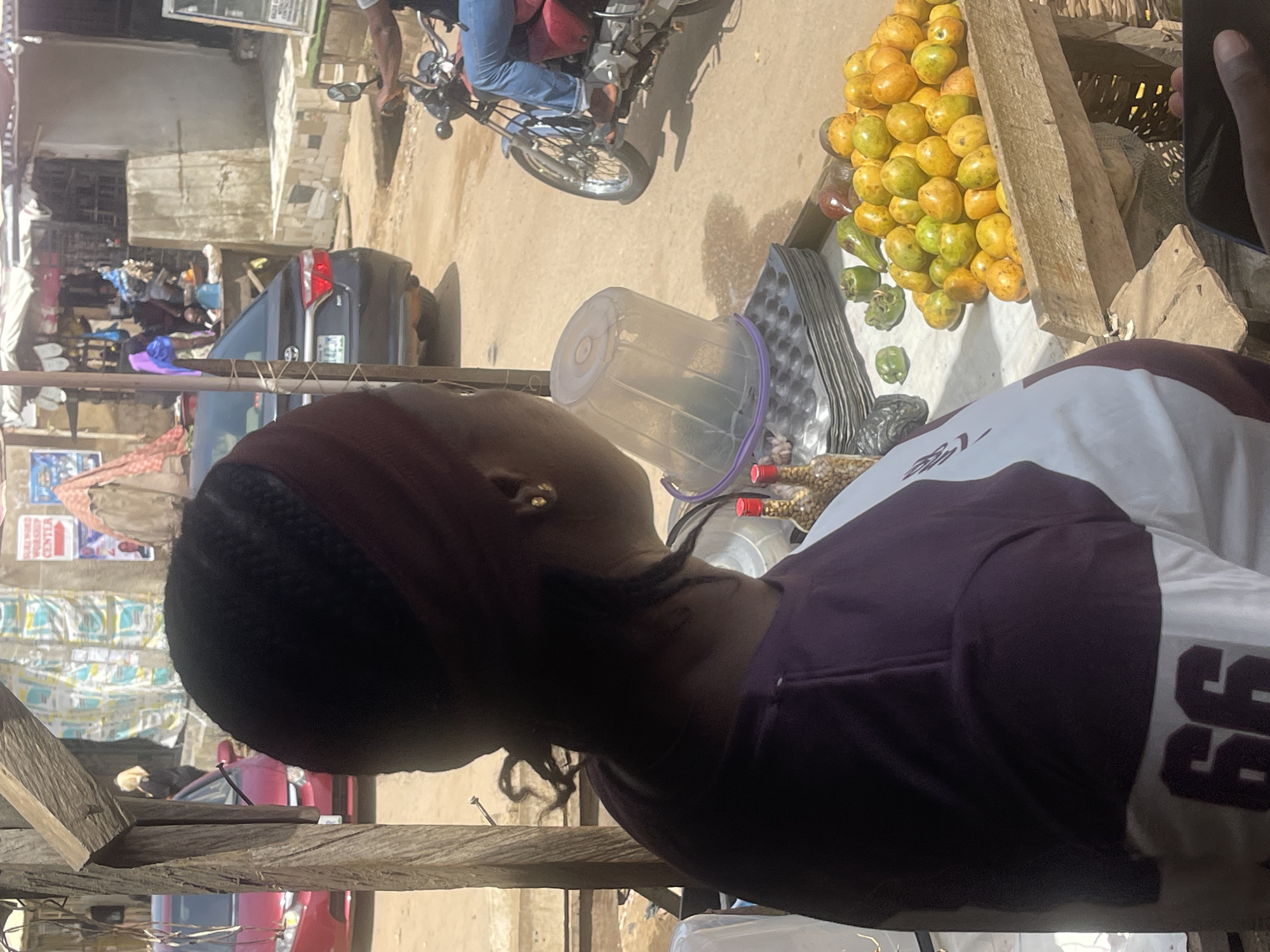 The image shows a vendor in a bustling street market in Abuja, Nigeria. The vendor, an African woman with a dark headband, is positioned behind a wooden stall laden with produce. Oranges are prominently displayed, alongside a plastic bucket and metal trays. In the background, a motorcycle is parked, and various vehicles and pedestrians move along the street. The scene suggests a busy and active marketplace environment.