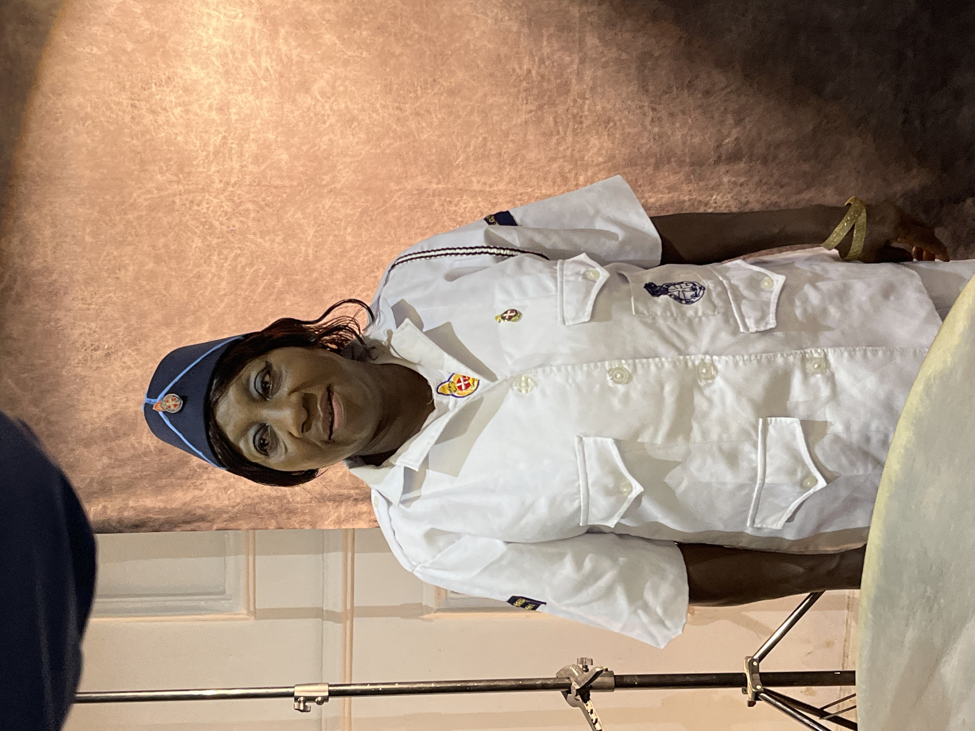 A medium shot shows a Black woman in a white uniform, including a cap, standing in front of a textured, copper-colored backdrop. The woman is looking towards the viewer with a slight smile. Her uniform has several pockets and appears to be a type of formal or ceremonial attire. She wears a dark blue cap with a red and blue emblem. There are also various insignias on her uniform, including one on her left breast and another on her left sleeve. The backdrop has an uneven, textured surface that is lighter at the top and darker at the bottom. To the left of the woman, part of a lighting stand is visible. The lighting appears to be coming from the front and slightly from the left, creating some shadows on the right side of the woman and the backdrop. In the foreground, a portion of a light-colored, curved object, possibly a drum, is visible.