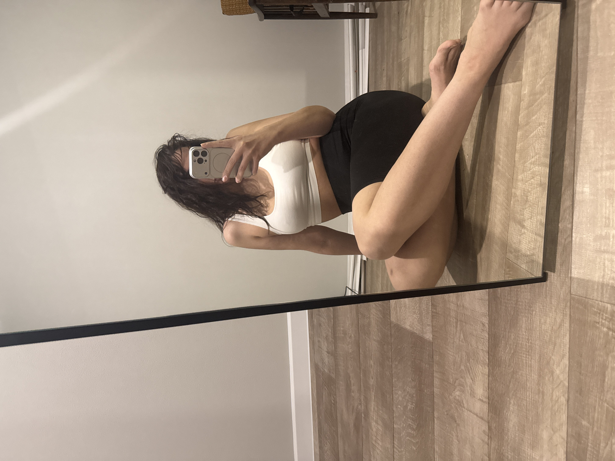 A young woman is posing for a selfie in a mirror. She is wearing a white sports bra and black shorts. She has dark, wavy hair. The room appears to be a bedroom with wood flooring and light-colored walls. There is a small woven basket visible behind her, possibly on top of a piece of furniture. The image does not provide location information; therefore, specifying Chaikovsky, Russia, is not possible.