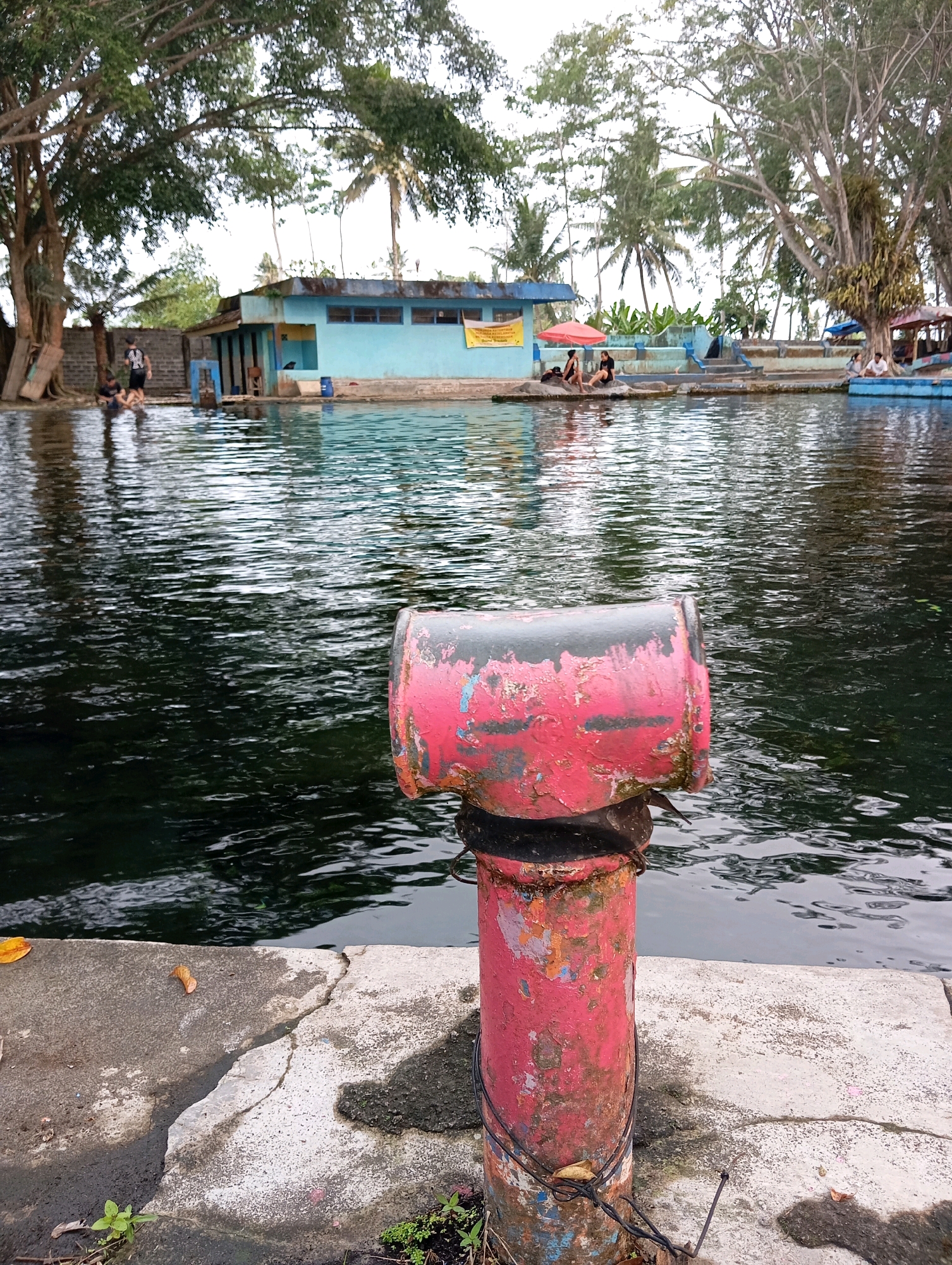A body of dark, rippling water is in the foreground of the image. The water is bordered by a concrete edge, which is cracked in some places. A rusty red and pink post with peeling paint stands in the foreground, to the right of the image center. The post appears to be a bollard. In the background, a light blue building with a yellow sign is visible. Several palm trees and other foliage are present around the building and extending into the background. There are people in the water and on the edge of the water. Two people sit on rocks in the mid-ground, and several people are visible in the water near the blue building. The sky is overcast. The location is identified as Sememu, Indonesia.