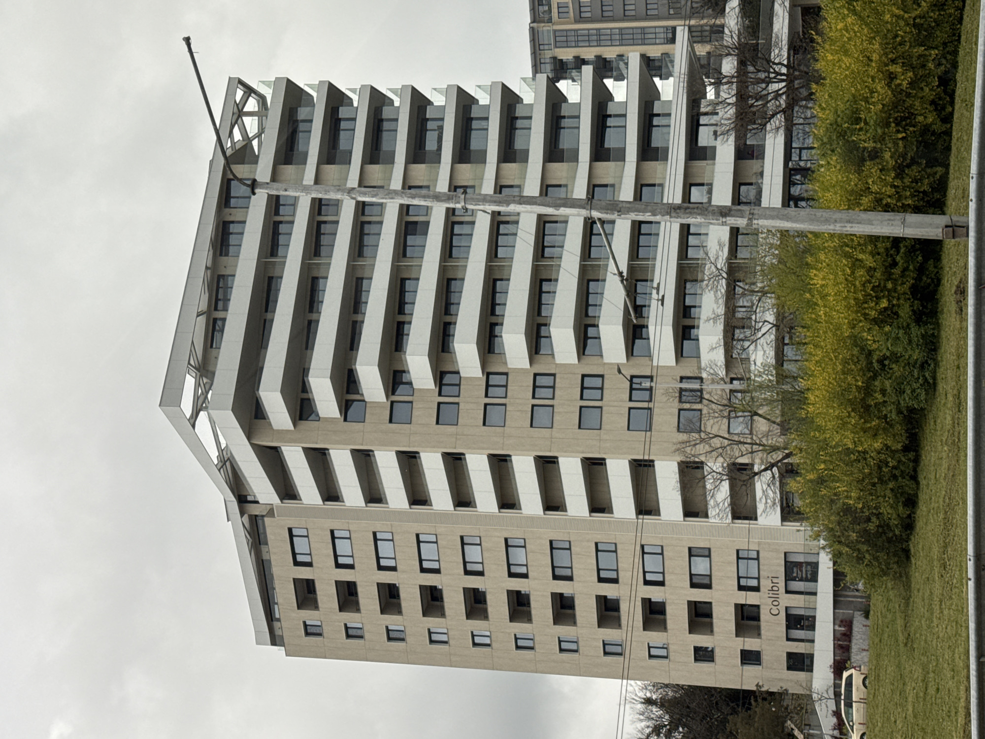 The image shows a multi-story building with a modern architectural design, likely residential or commercial, against a cloudy sky. The building has a repeating pattern of windows and projecting white structural elements. A utility pole is visible in the foreground, running vertically through the frame. To the right, green foliage suggests a park or landscaped area. The name "Colibri" is visible on the building, indicating a possible business or location name. Based on architectural style and the "Colibri" signage, the building is likely located in the city of Varna, Bulgaria.