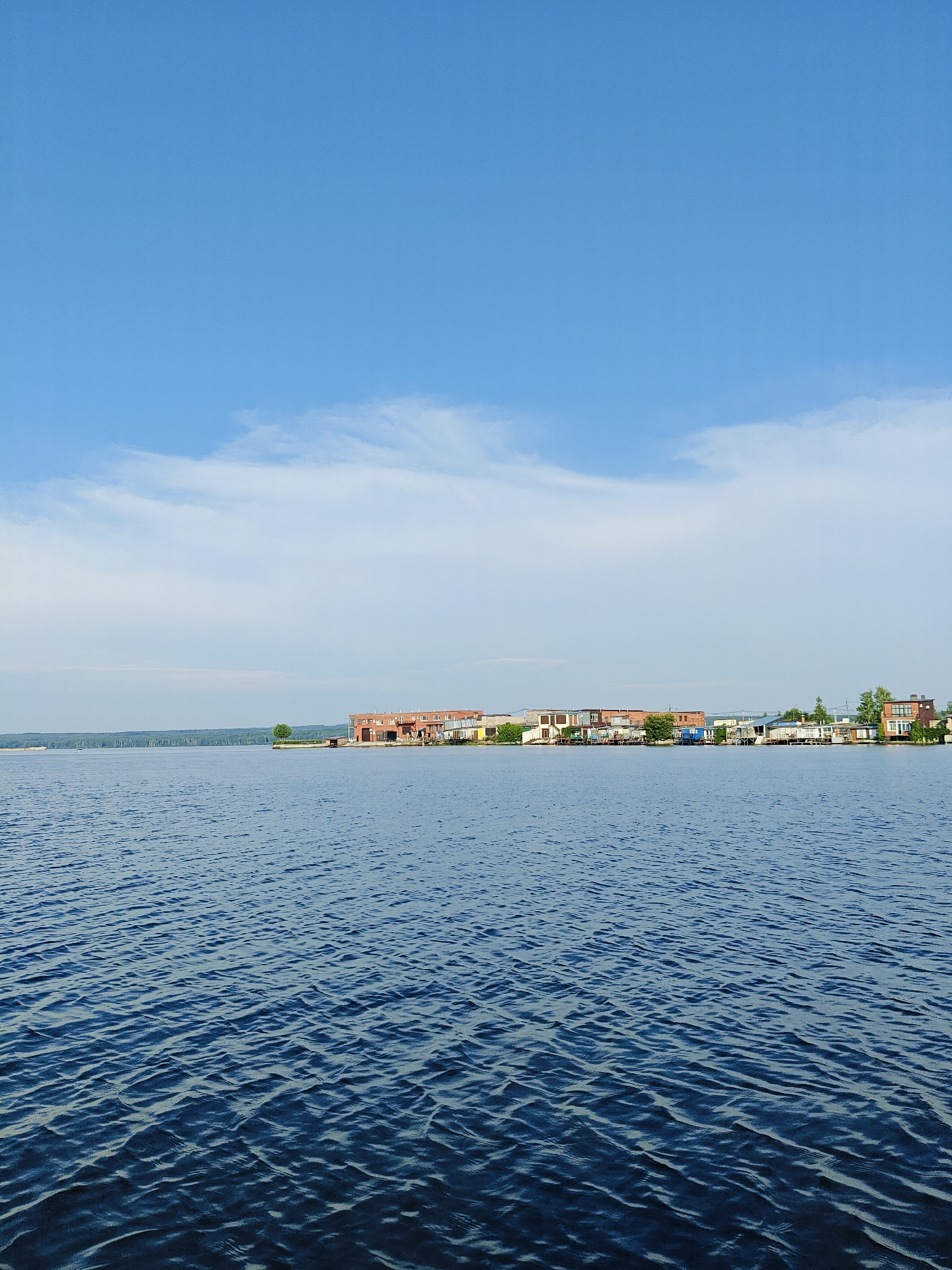 A wide, tranquil blue lake stretches across the lower half of the image, with gentle waves rippling on its surface. In the distance, along the far shore, a line of buildings is visible, appearing as a residential or industrial area. The buildings vary in color and style, with some having brick facades and others appearing more modern. A bridge or causeway extends across the water to the left, connecting to the distant shore. The sky above is a clear, bright blue with a few wispy white clouds scattered across it. The overall scene suggests a calm day in a lakeside or riverside community.