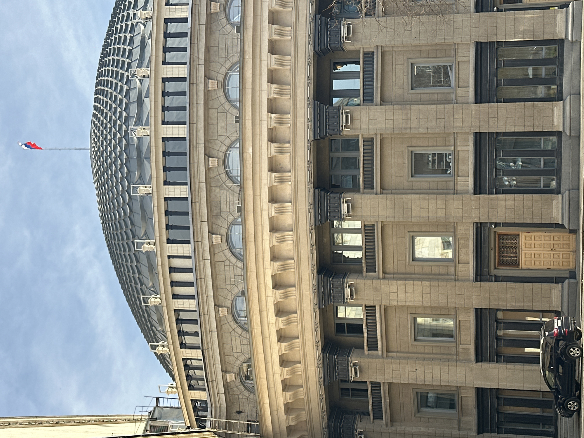 The image depicts a section of a building in Novosibirsk, Russia. The structure exhibits architectural details including a domed roof with a patterned surface, windows, and decorative elements. A flag is visible atop the dome. A car is parked in front of the building. The image seems to be taken from a low angle.