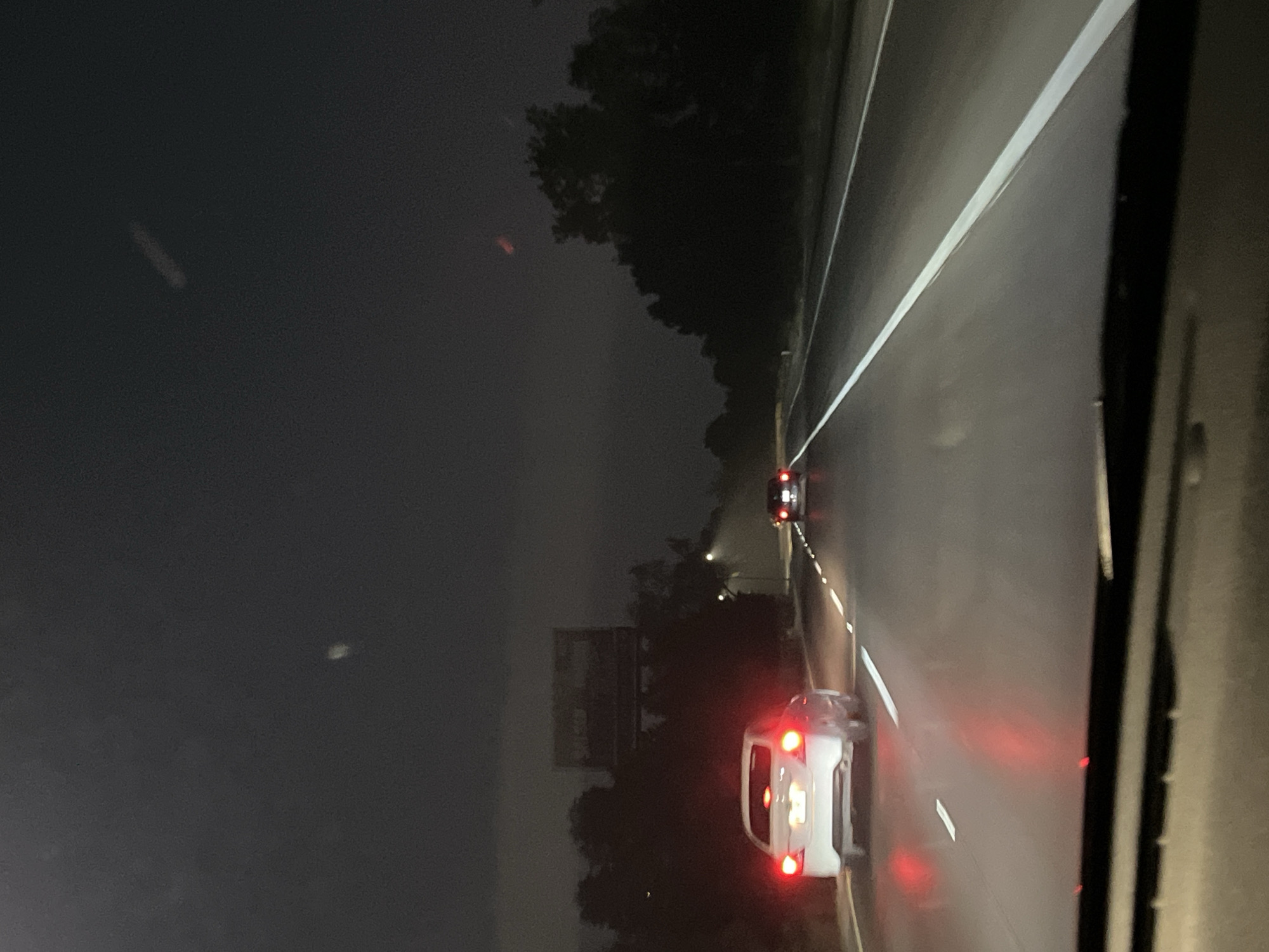 A nighttime photograph depicts a highway scene, likely taken from the perspective of the driver of a car. Two vehicles with illuminated taillights are visible on the road ahead. A billboard is located on the side of the highway. The environment is dark with trees lining the highway. Barrio de Nuevo León is not discernible from the image.