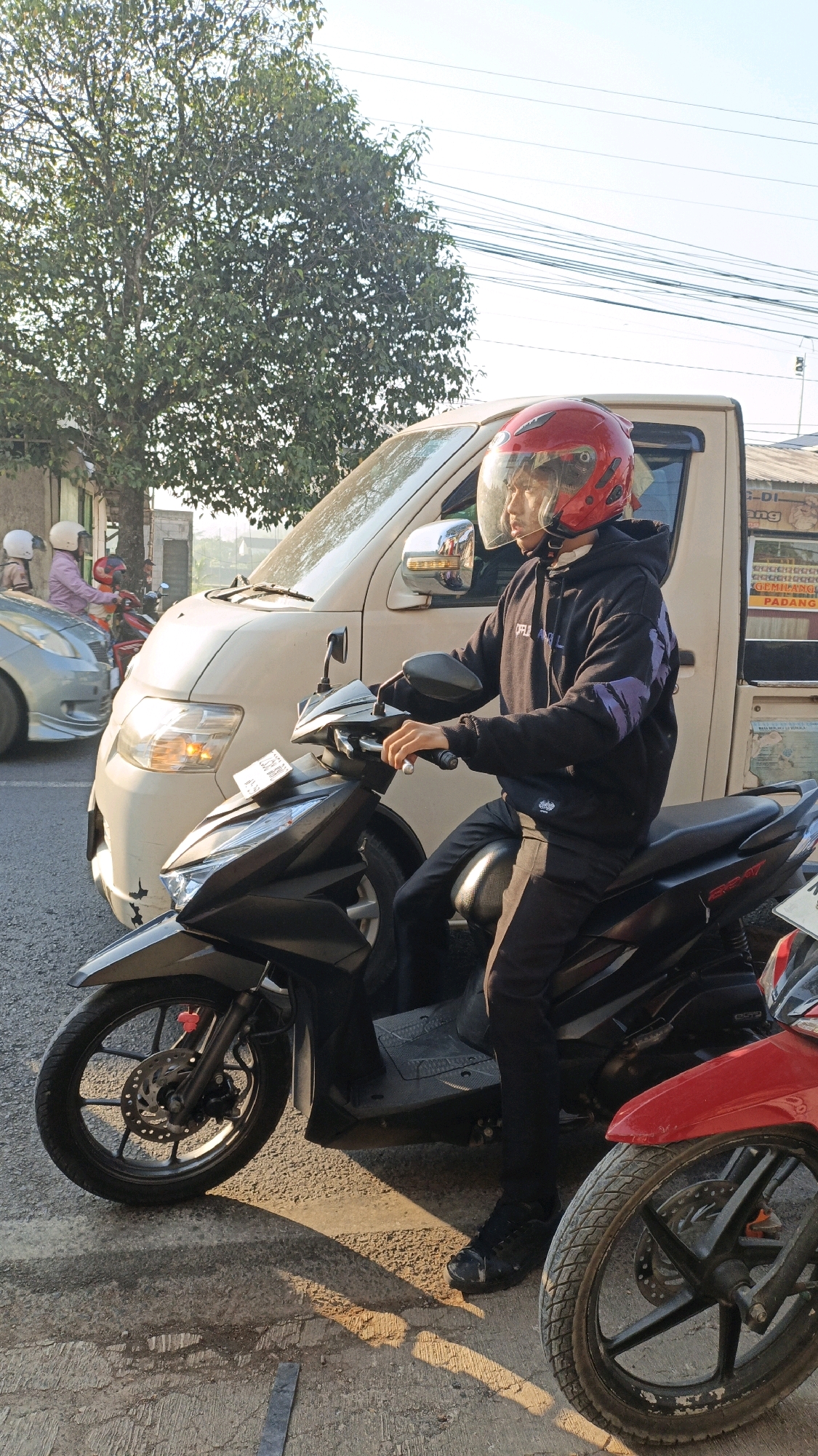 A person wearing a red helmet and a black hoodie with purple accents is sitting on a black scooter. They are on a street in Kuwasen, Indonesia, next to a light-colored van. The street is paved and appears to be dry. There is a tree with green leaves to the left of the scene and other vehicles and buildings in the background. The lighting suggests it is either early morning or late afternoon, with long shadows cast on the ground. The scooter has a license plate.